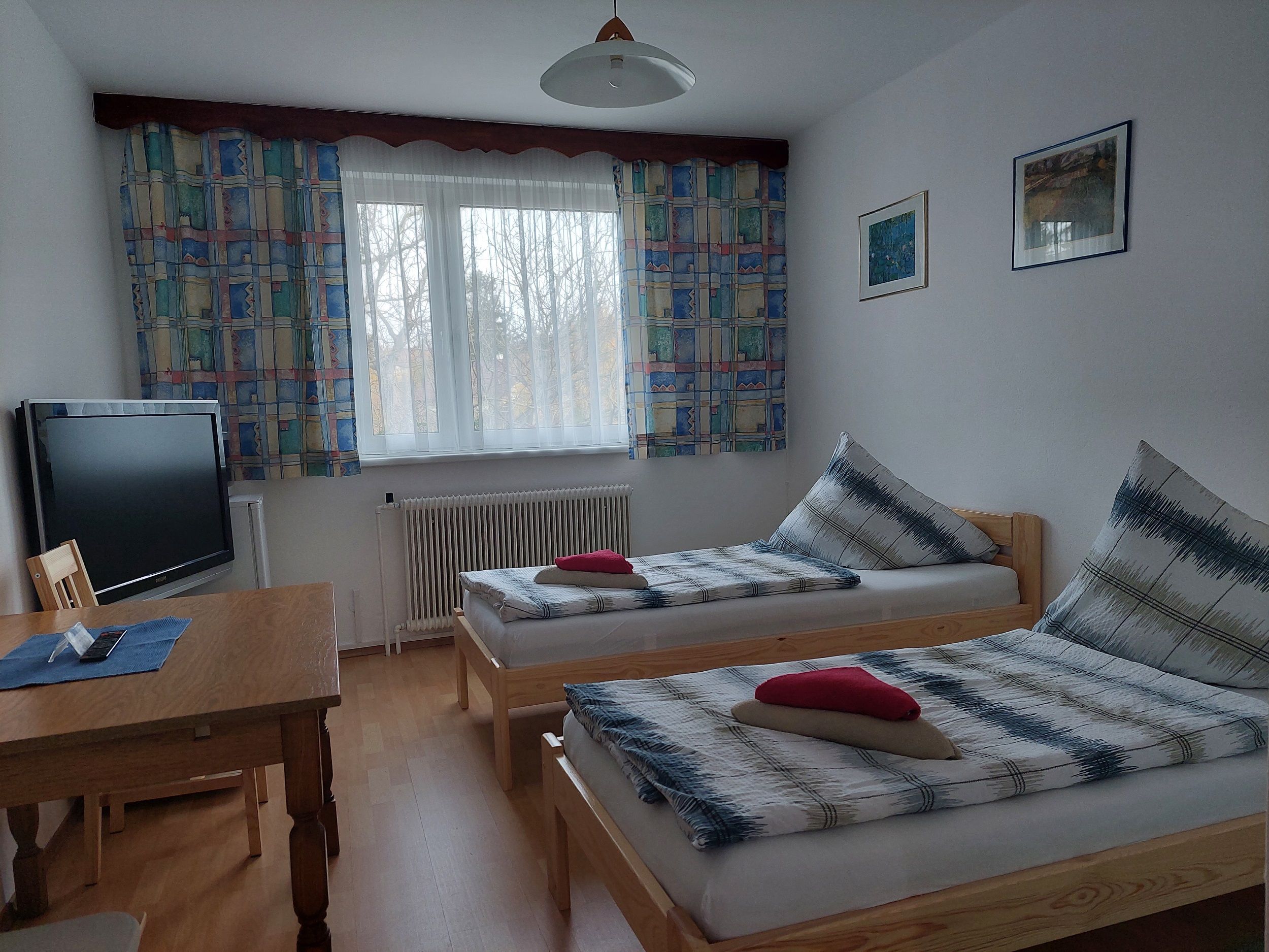 Egyszerűen berendezett szoba két egyszemélyes ággyal, asztallal, TV-vel és színes függönyökkel ellátott ablakkal.