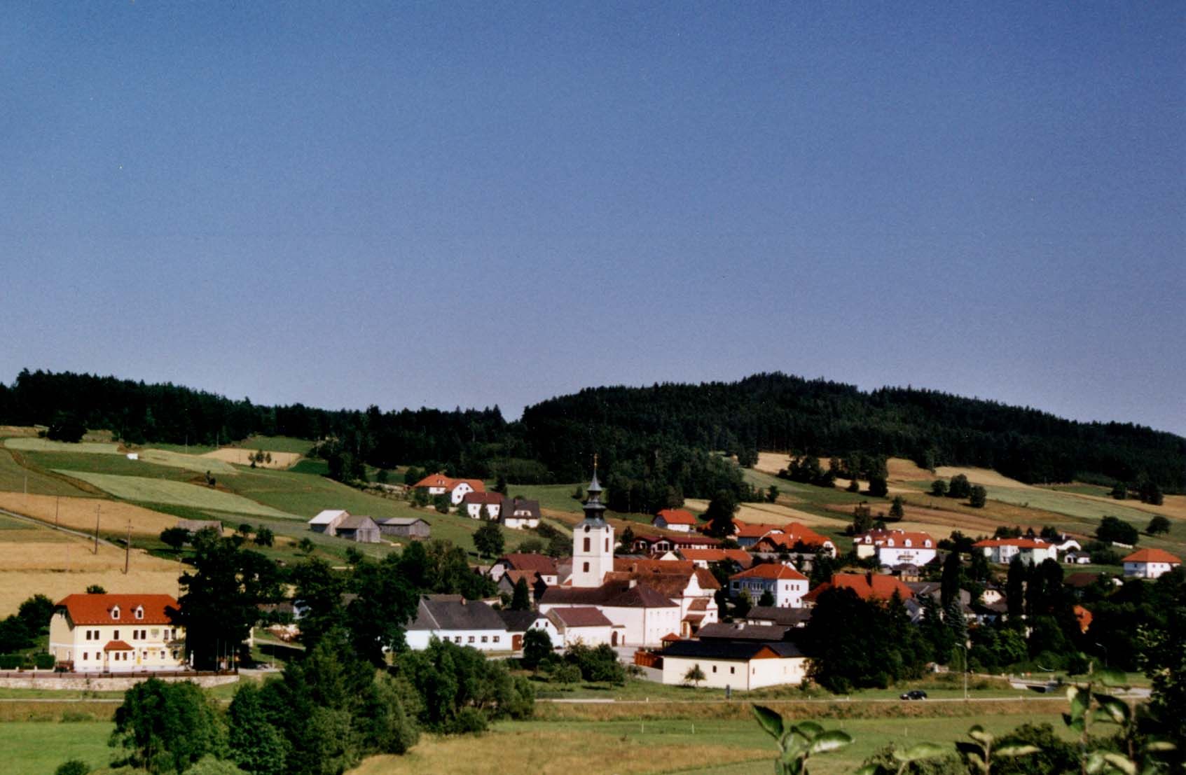 Panoráma egy templomos, piros tetős falura, amelyet zöld mezők és erdők vesznek körül.