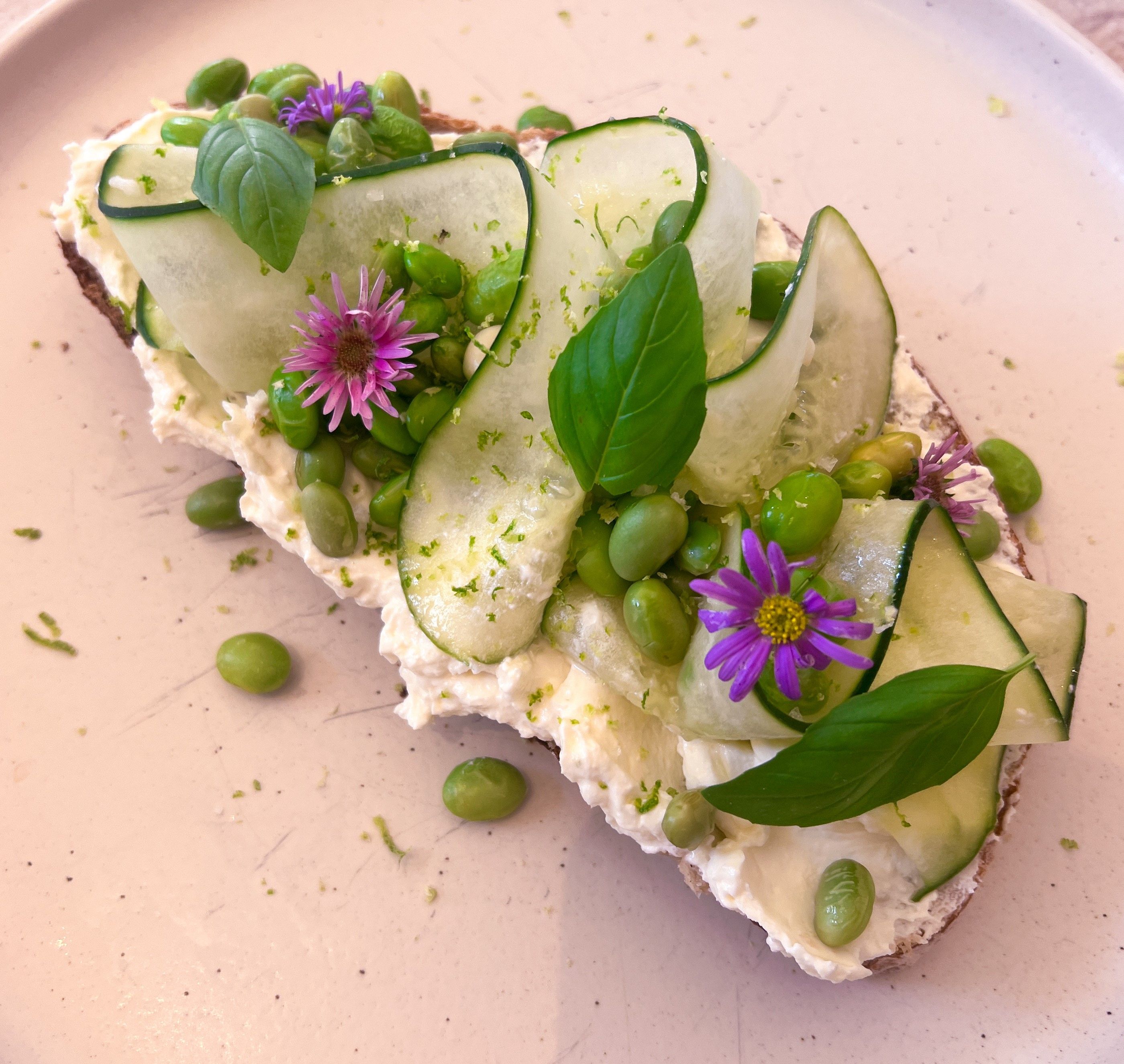 Egy szendvics krémsajttal, uborkaszeletekkel, edamame-mal, bazsalikomlevelekkel és lila virágokkal egy tányéron.
