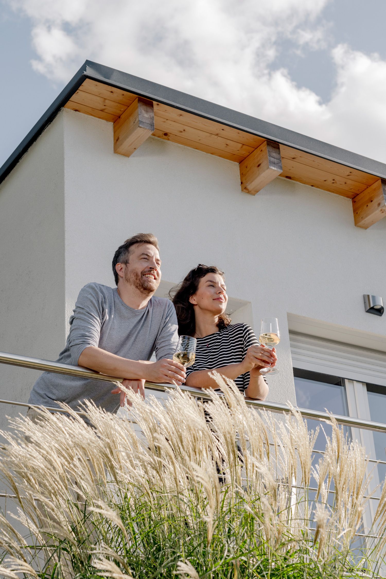 Egy pár áll egy erkélyen, kezében borospohárral, a háttérben egy modern épület.