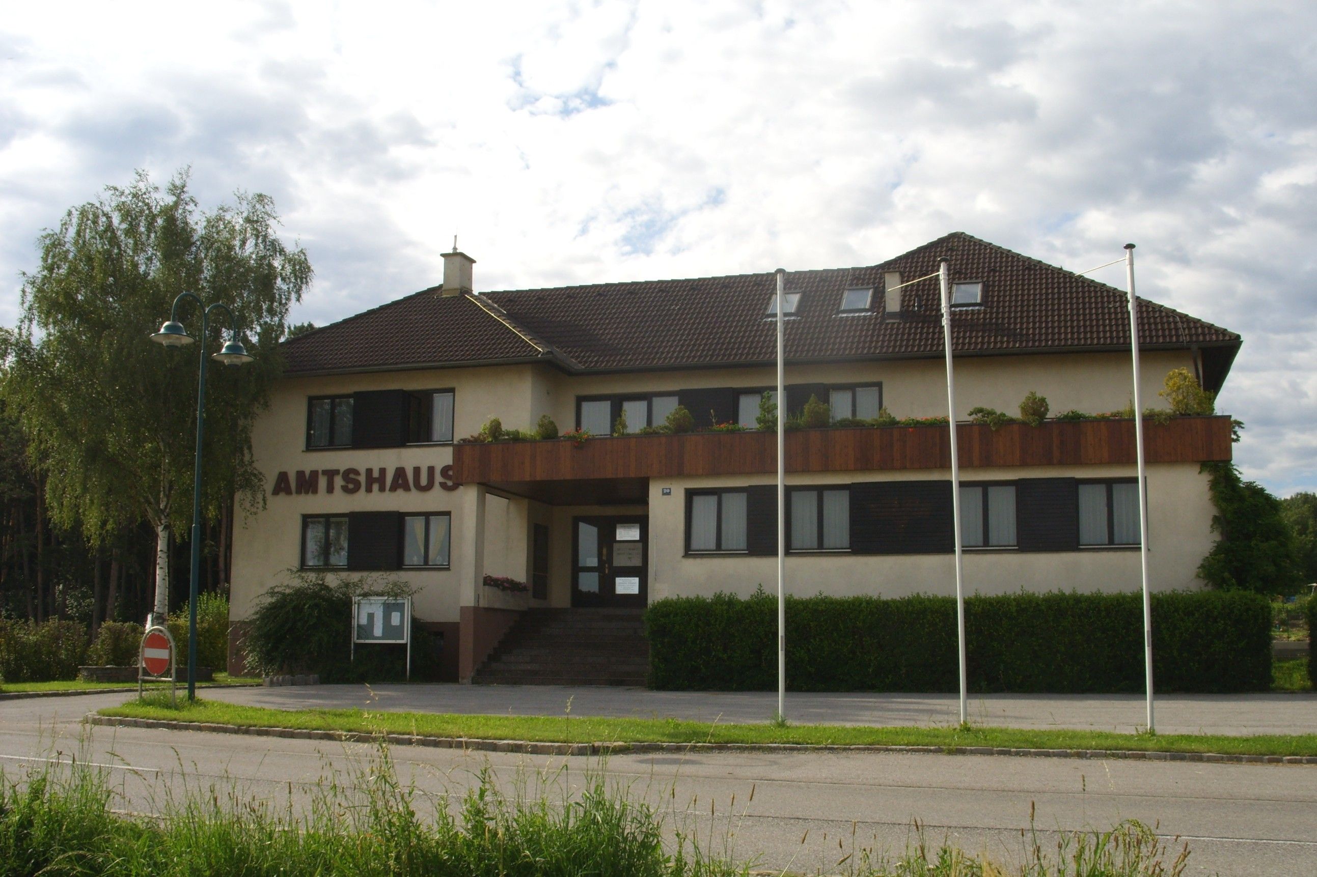 A Natschbach-Loipersbach irodaház barna tetővel és erkéllyel, fákkal és zászlórudakkal körülvéve.