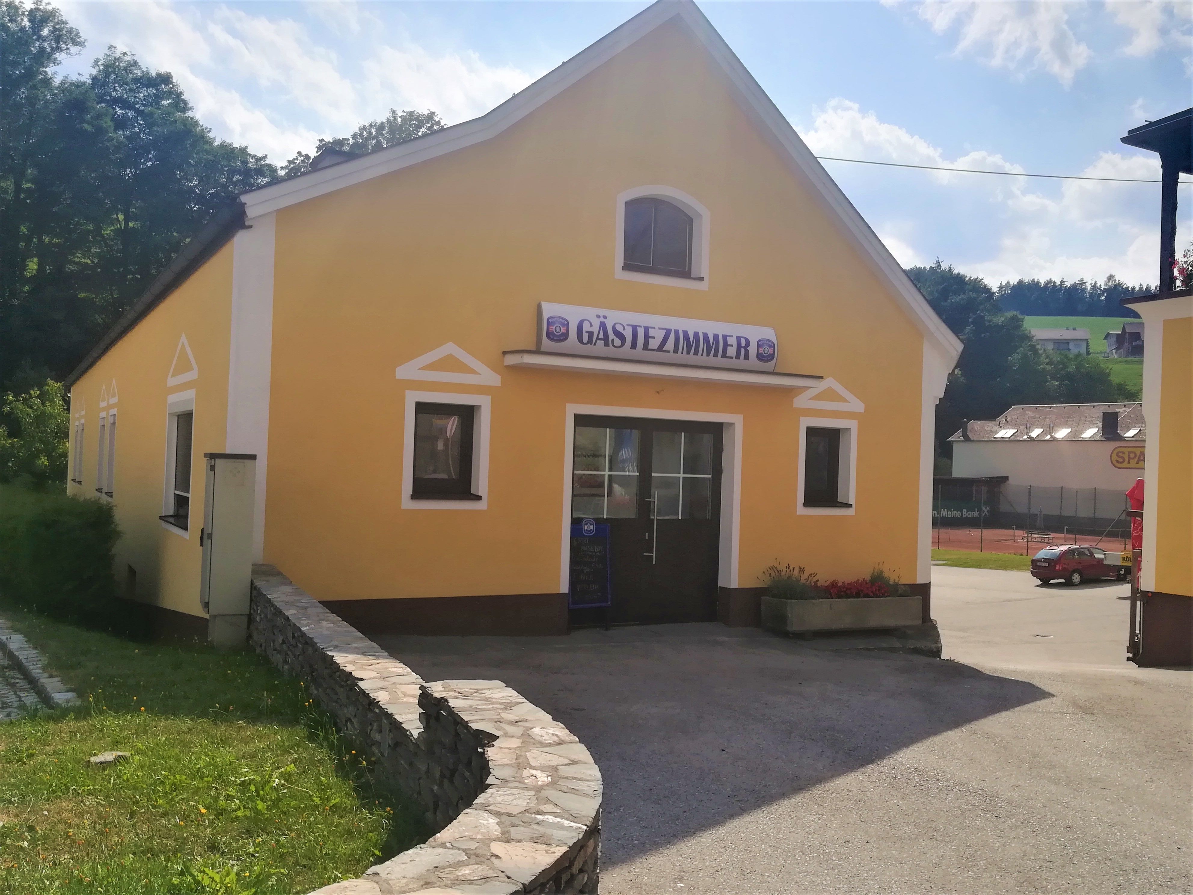 Sárga épület "vendégszoba" felirattal, fákkal és parkolóval körülvéve.