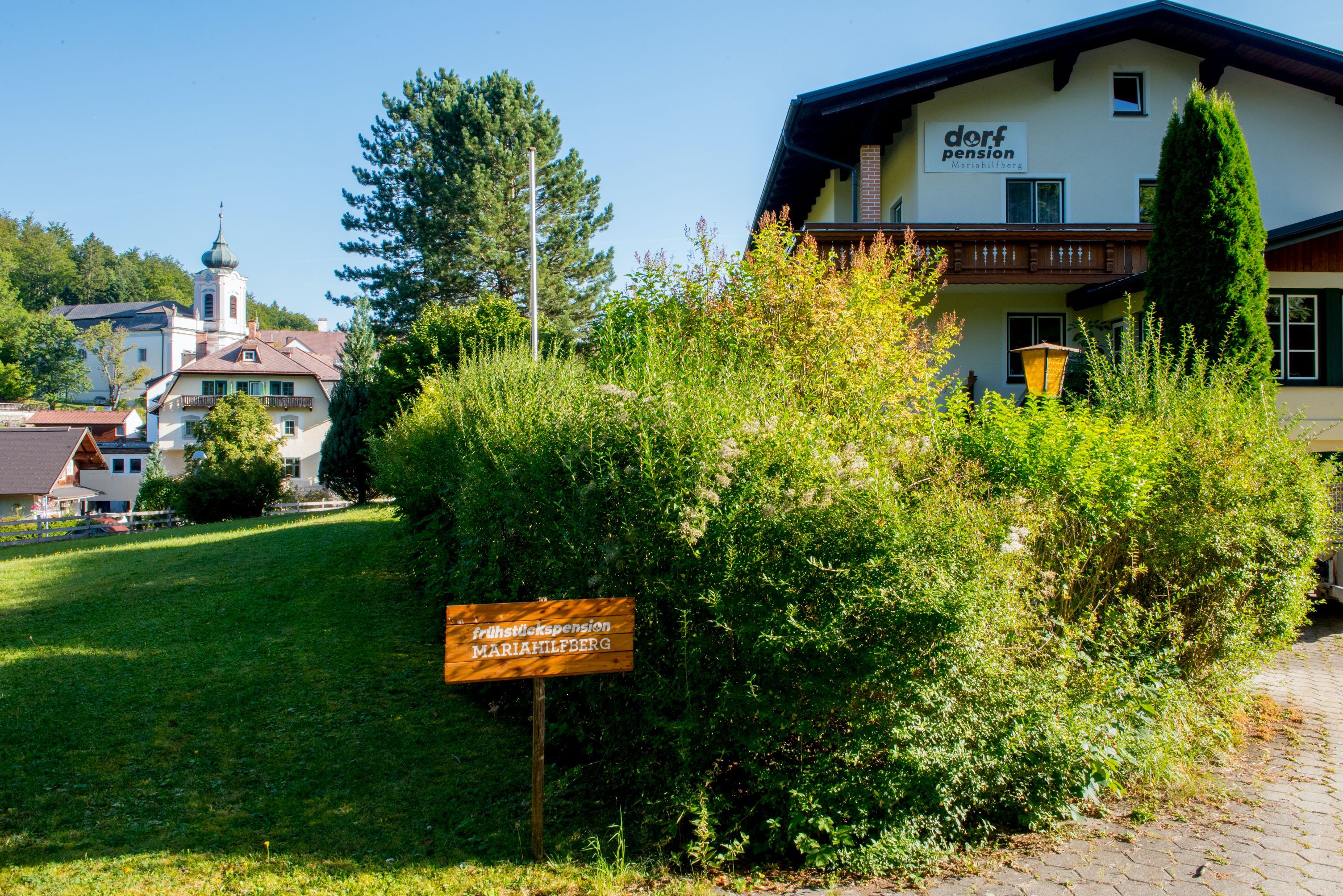 Egy "Dorfpension" feliratú épület és egy "Frühstückspension Mariahilfberg" feliratú tábla zöld környezetben.