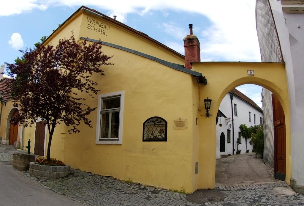 Sárga épület "Weinbau Schabl" felirattal, mellette egy boltív és egy fa.