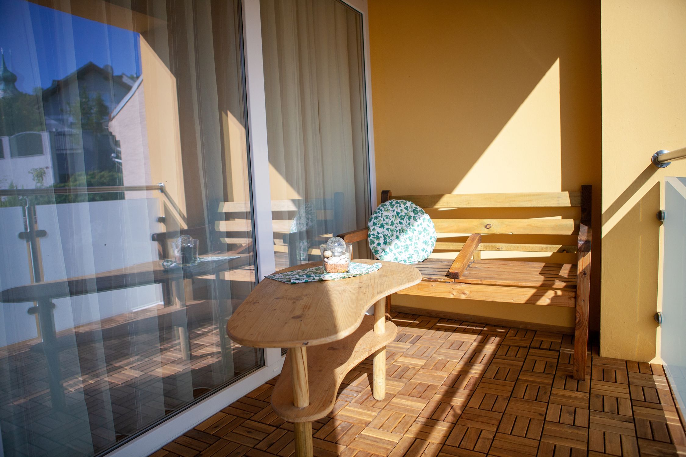 Egy erkély fapadlóval, fa asztallal és egy kerek párnával ellátott fapaddal. A háttérben egy üvegkorlát és egy épület látható.