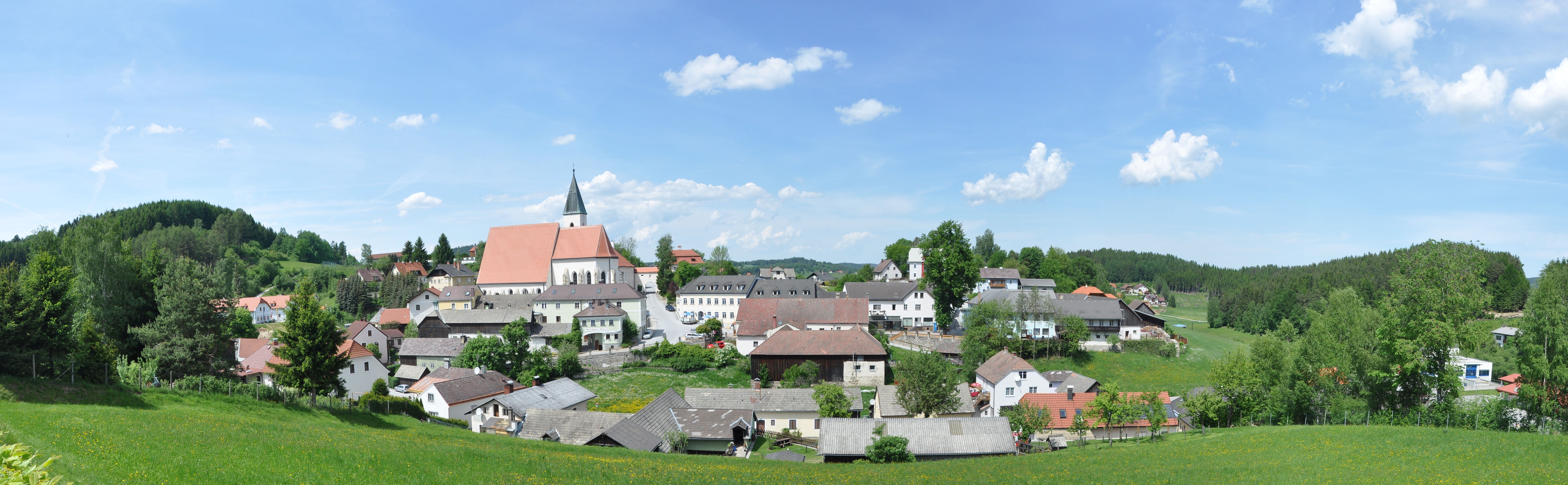 Schönbach mezővárosának panorámája a templommal és a környező házakkal, zöld rétekkel és erdőkkel körülvéve, kék ég alatt.