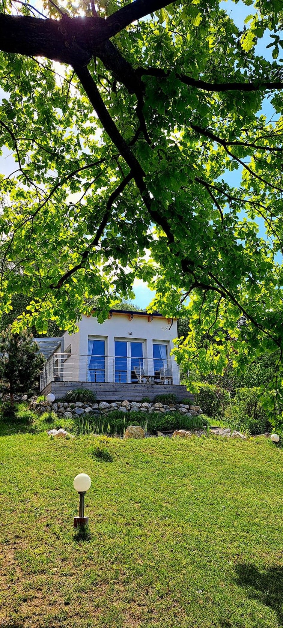 Egy modern faház nagy ablakokkal, zöld természettel és fákkal körülvéve.