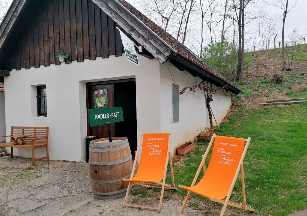 Egy kis fehér épület, fatetővel, két narancssárga nyugágydal és egy fahordóval előtte, fákkal és fűvel körülvéve.
