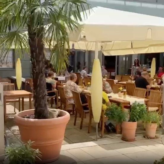 Egy kávézó terasza, faasztalokkal és -székekkel, növényekkel körülvéve és egy nagy napernyővel.
