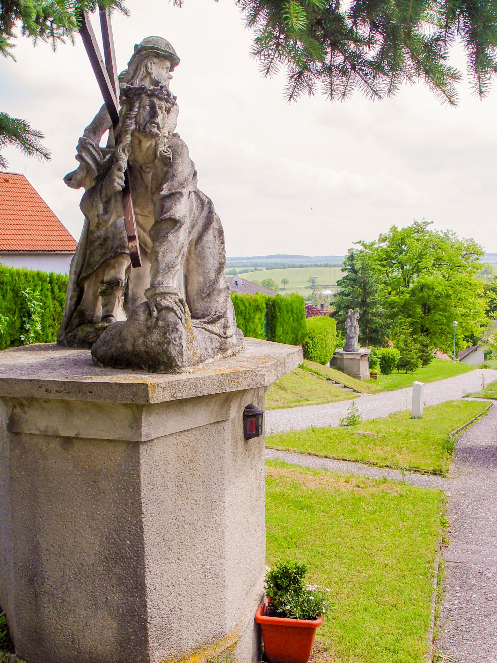 Kőszobor talapzaton, háttérben zöld táj.