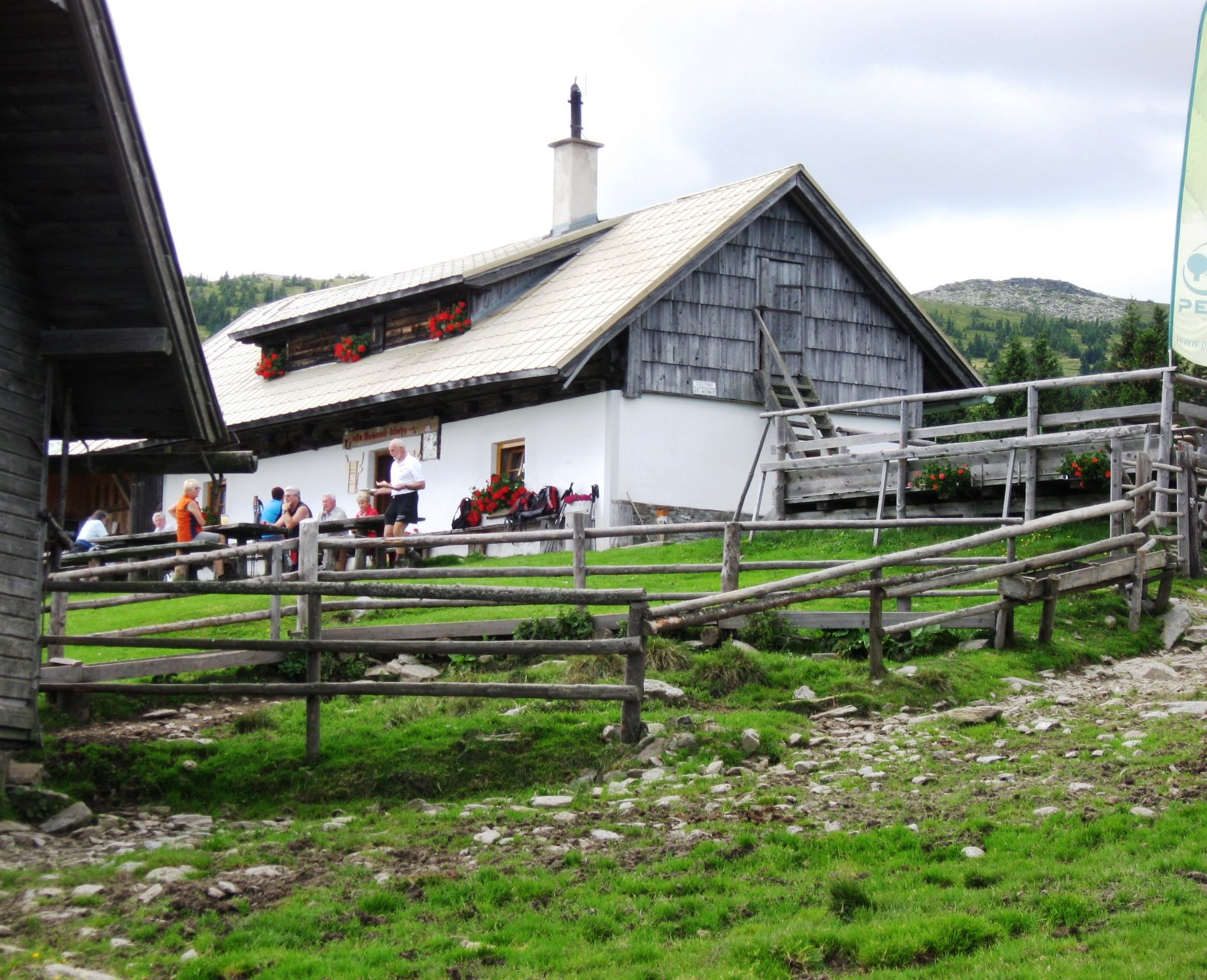 Alpesi hütte emberekkel egy réten, fakerítéssel körülvéve.