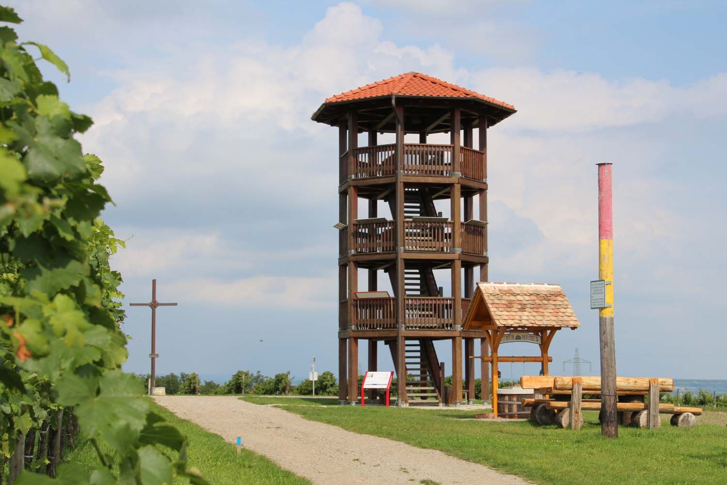 Egy vörös tetejű, fából készült kilátótorony áll egy dombon egy kereszt és egy pad mellett. Az ég felhős.