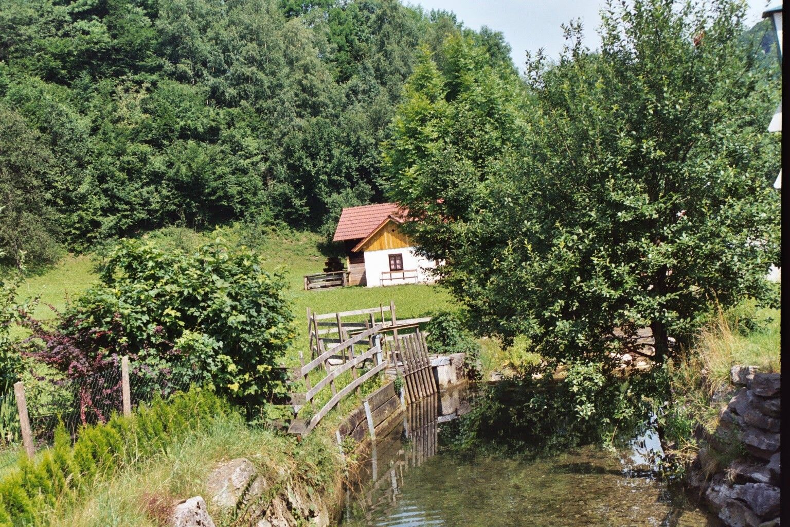 Egy kis piros tetejű turistaház áll a zöld tájban, egy patak és fák mellett.