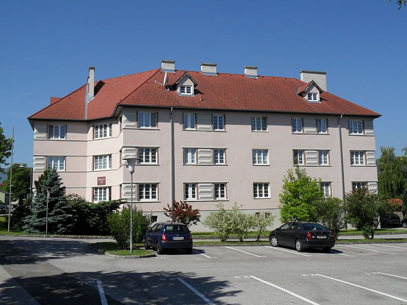 Lakóépület piros tetővel és több ablakkal, fákkal és parkoló autókkal körülvéve.