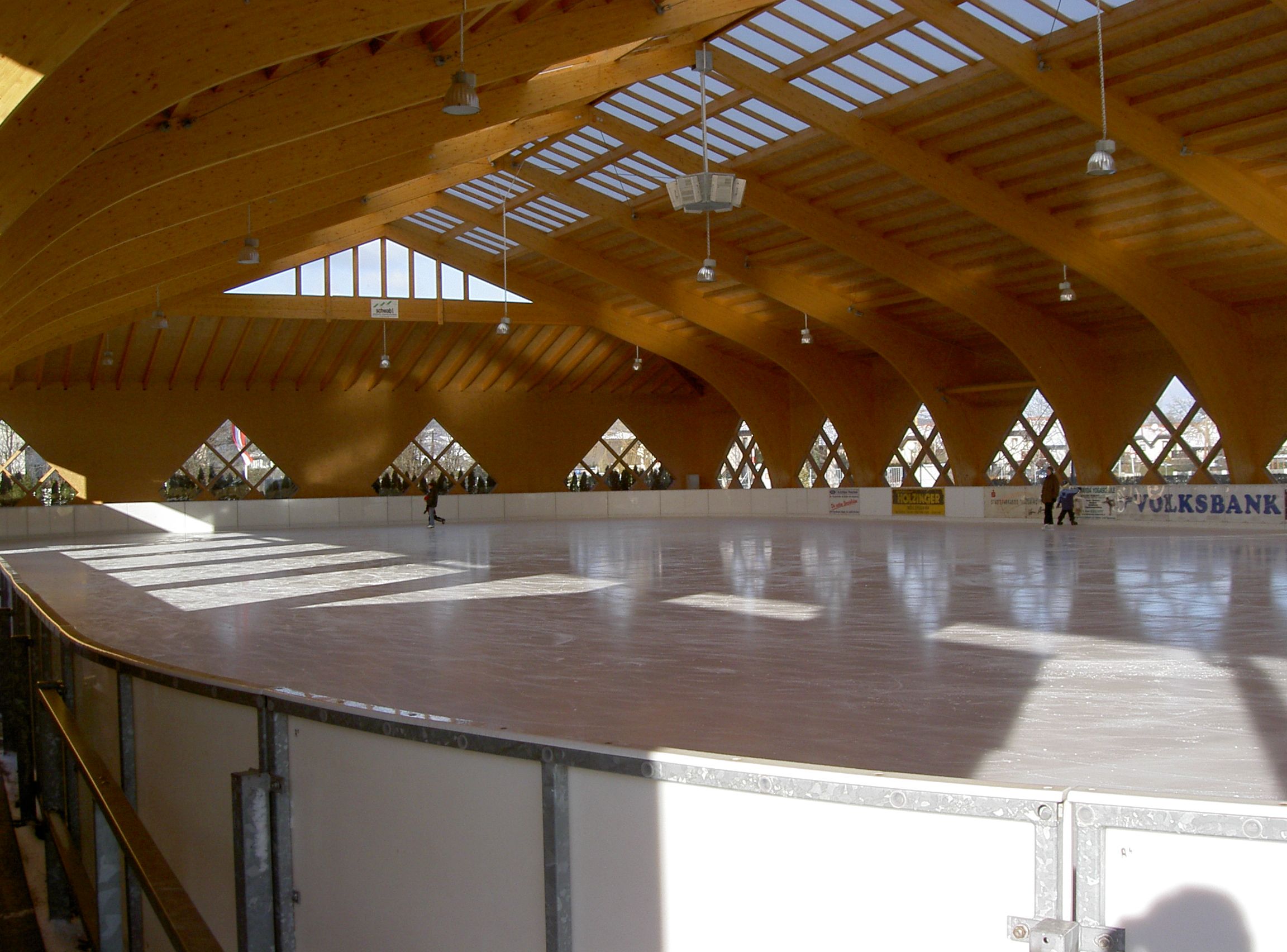 Egy fedett műjégpálya belső nézete, fából készült tetővel és kevés emberrel a jégen.