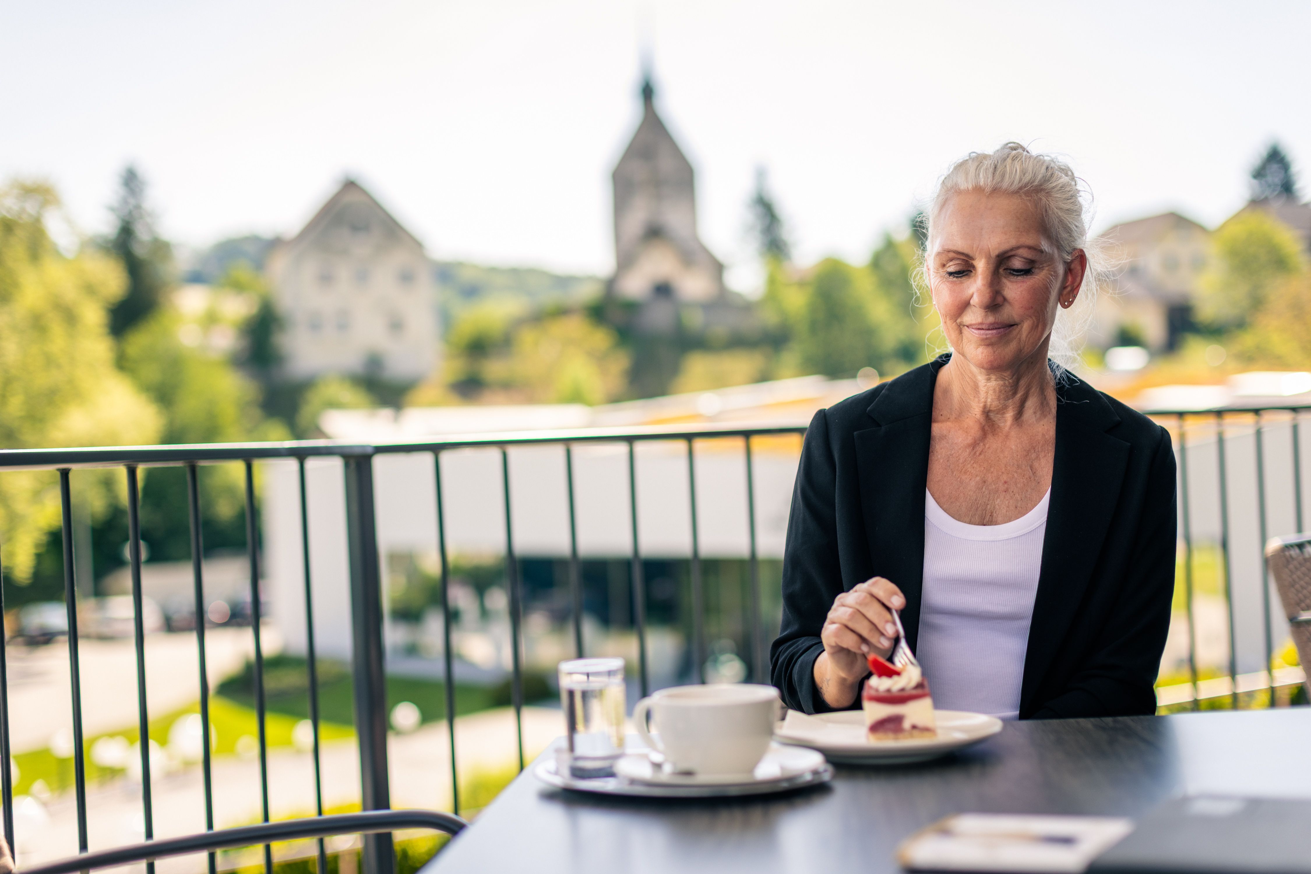 Egy nő ül a teraszon, és egy szelet tortát eszik egy csésze kávé mellett. A háttérben a Bad Schönau-i védőtemplom látható.