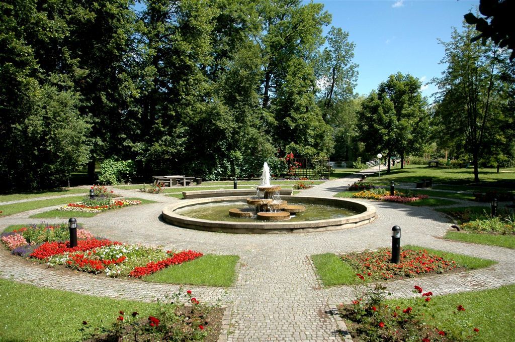Gondozott park, központi szökőkúttal, virágágyásokkal és fákkal körülvéve.