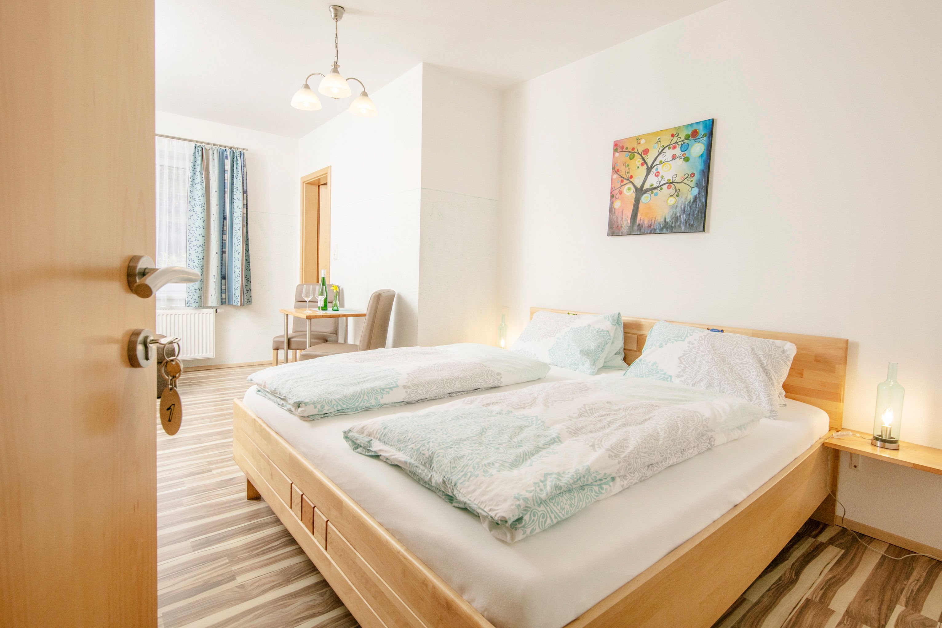 Világos vendégszoba franciaággyal, asztallal, székekkel és színes freskóval.