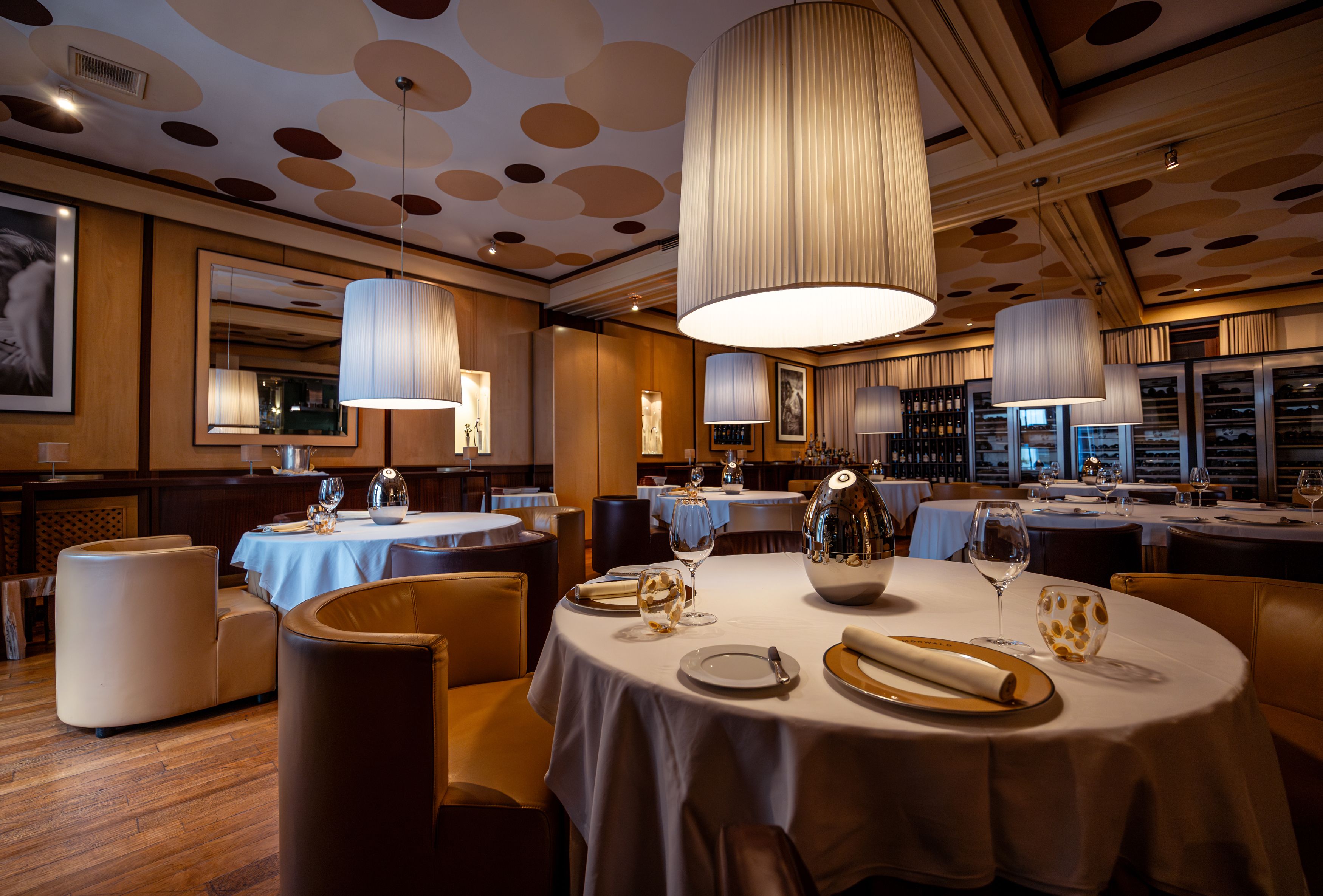 Elegáns étterem kerek asztalokkal, fehér abrosszal és nagy, lógó lámpákkal.
