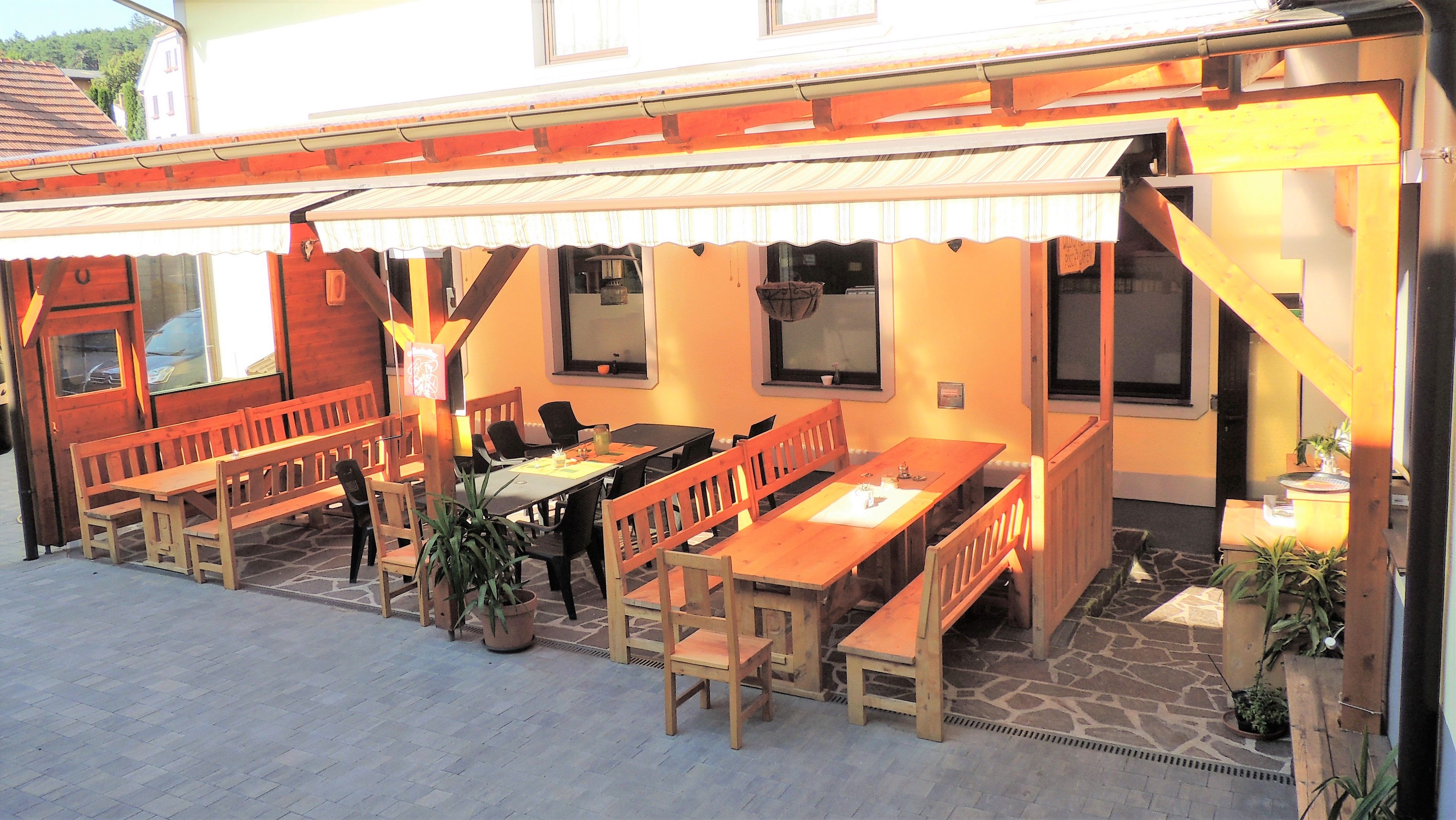 Hangulatos vendégkert fapadokkal és asztalokkal egy napellenző alatt.