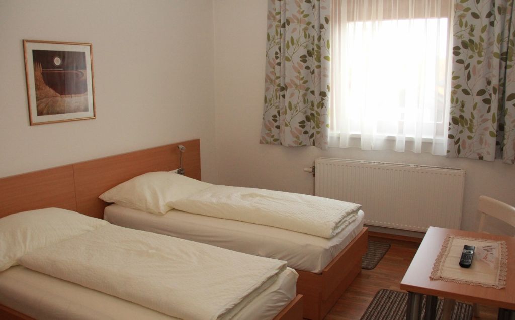 Vendégszoba két egyszemélyes ággyal, függönyös ablakkal, asztallal, távirányítóval és egy képpel a falon.