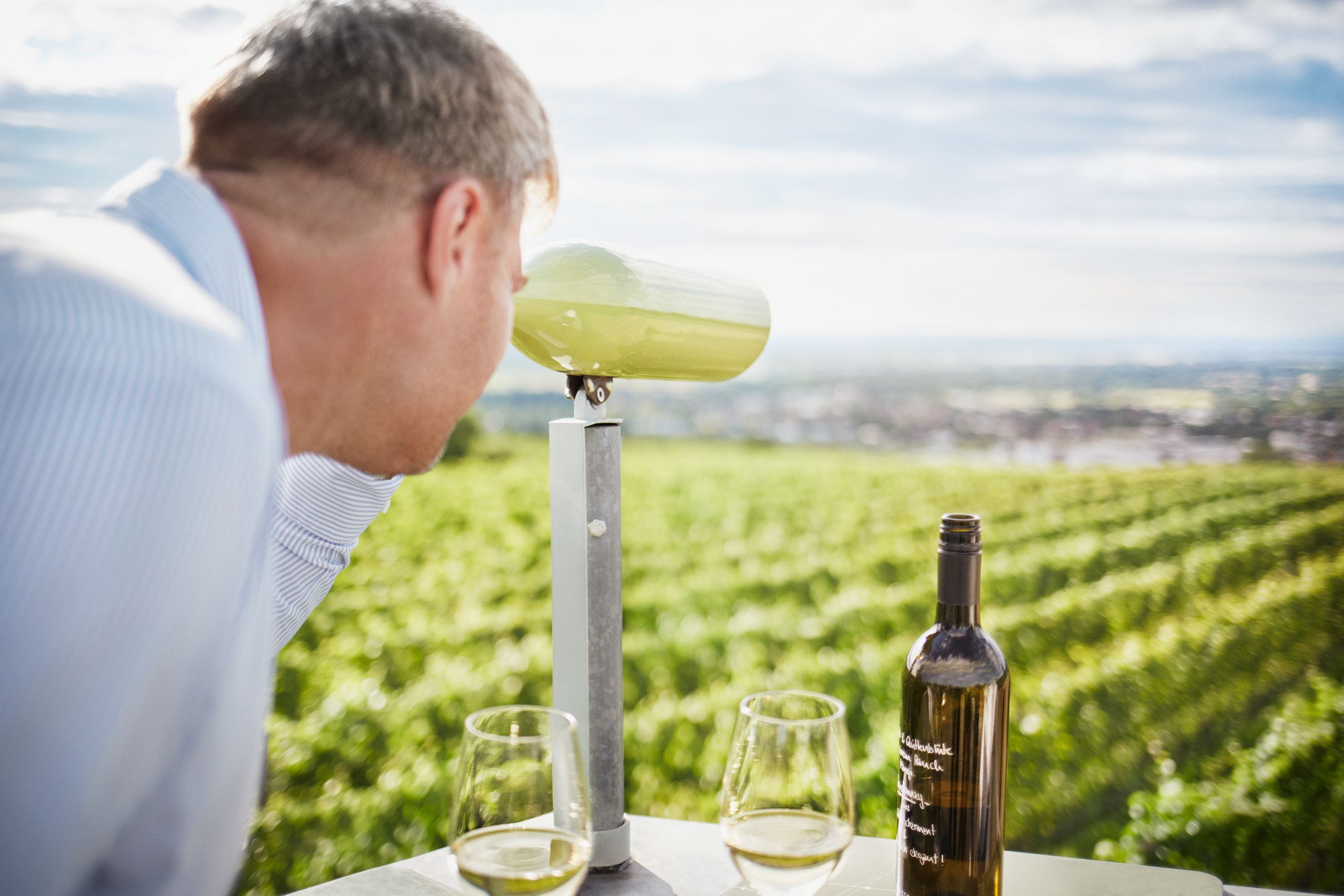 Egy férfi távcsövön keresztül nézi a szőlőmezőt. Az asztalon egy üveg bor és két pohár áll.