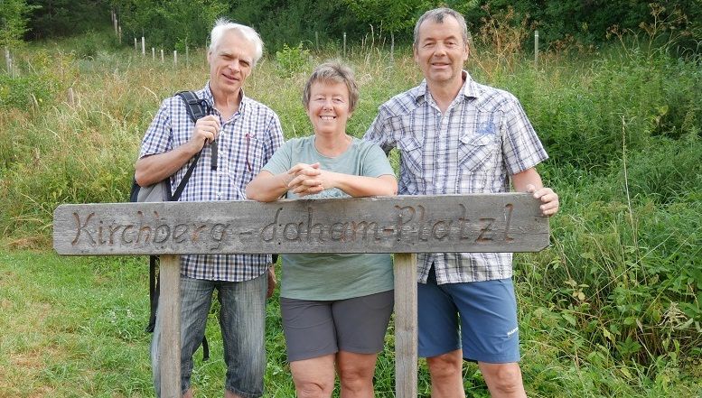 Három ember áll egy fatábla mögött, amelyen a "Kirchberg-daham-Platzl" felirat olvasható.