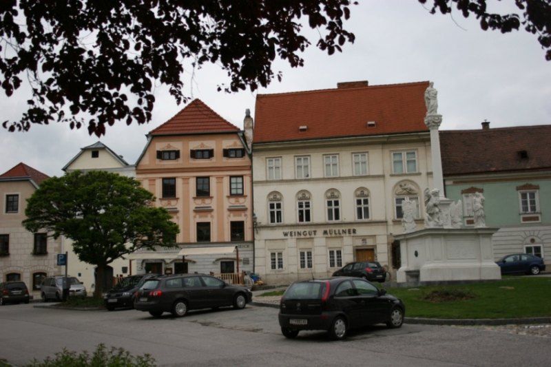 Történelmi épületek a "Weingut Müllner" felirattal és egy szoborral a téren.