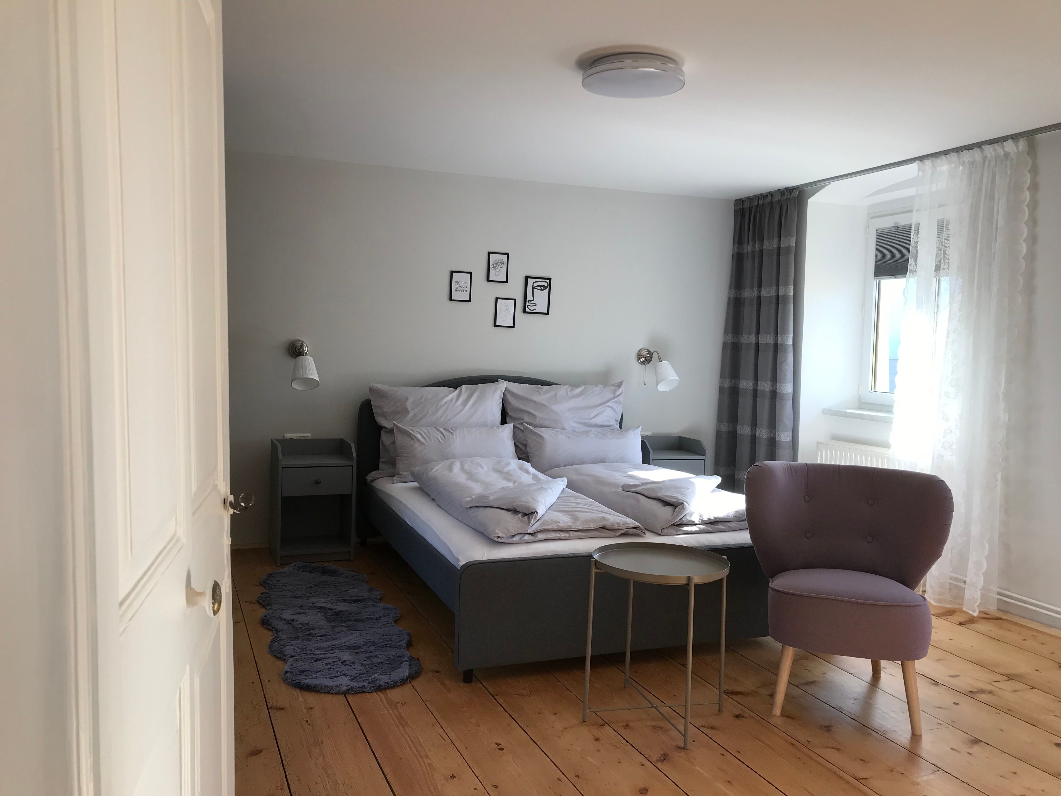 Modern bútorozott kétágyas szoba ággyal, karosszékkel és kis asztallal, fapadlón.