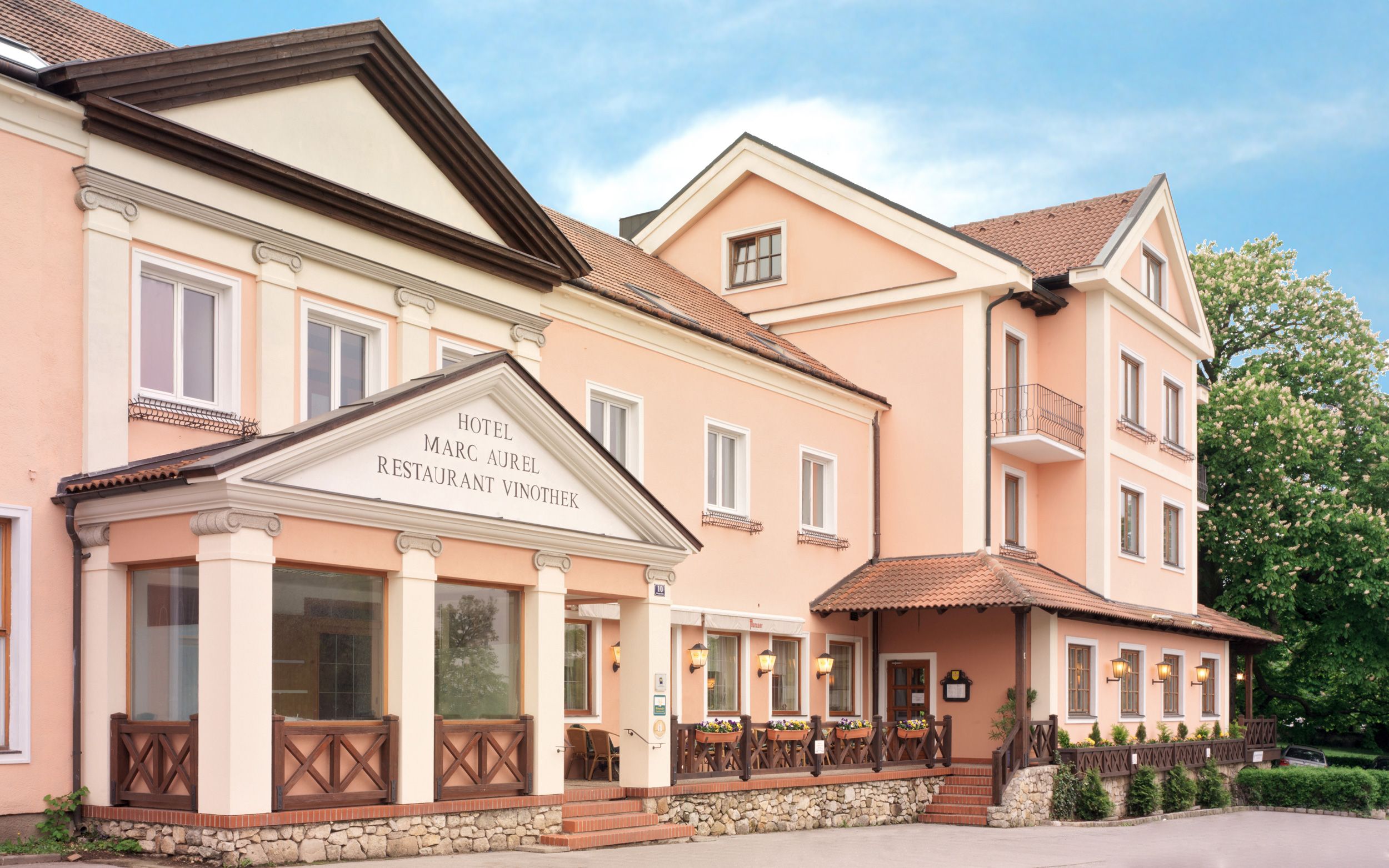 A Petronell-Carnuntumban található Hotel Marc Aurel külső nézete rózsaszín homlokzattal és klasszikus bejárattal.