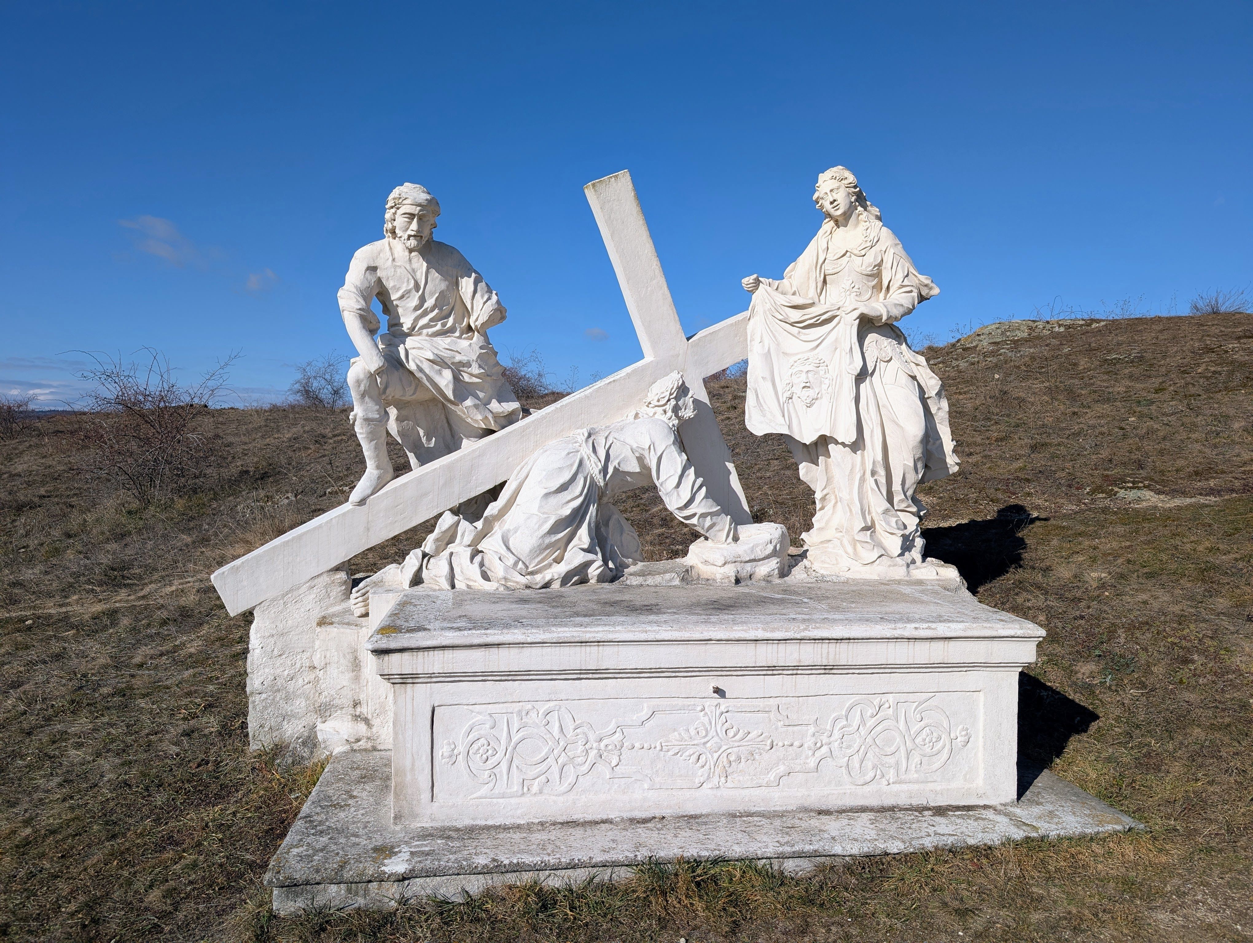 Vallási jelenetet ábrázoló kőszobor három alakkal és egy dombon álló kereszttel.
