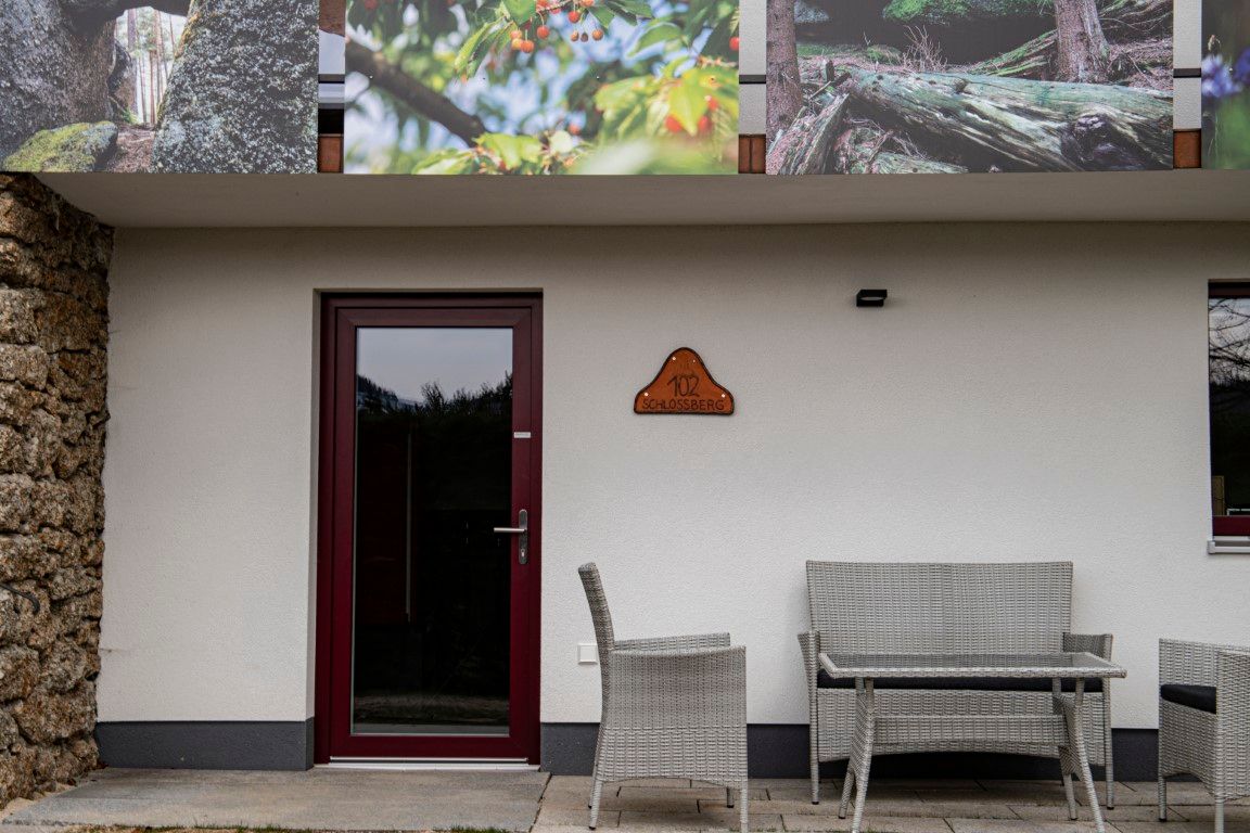 Egy épület bejárata, ajtóval, "102 Schlossberg" feliratú táblával és előtte kerti bútorokkal.