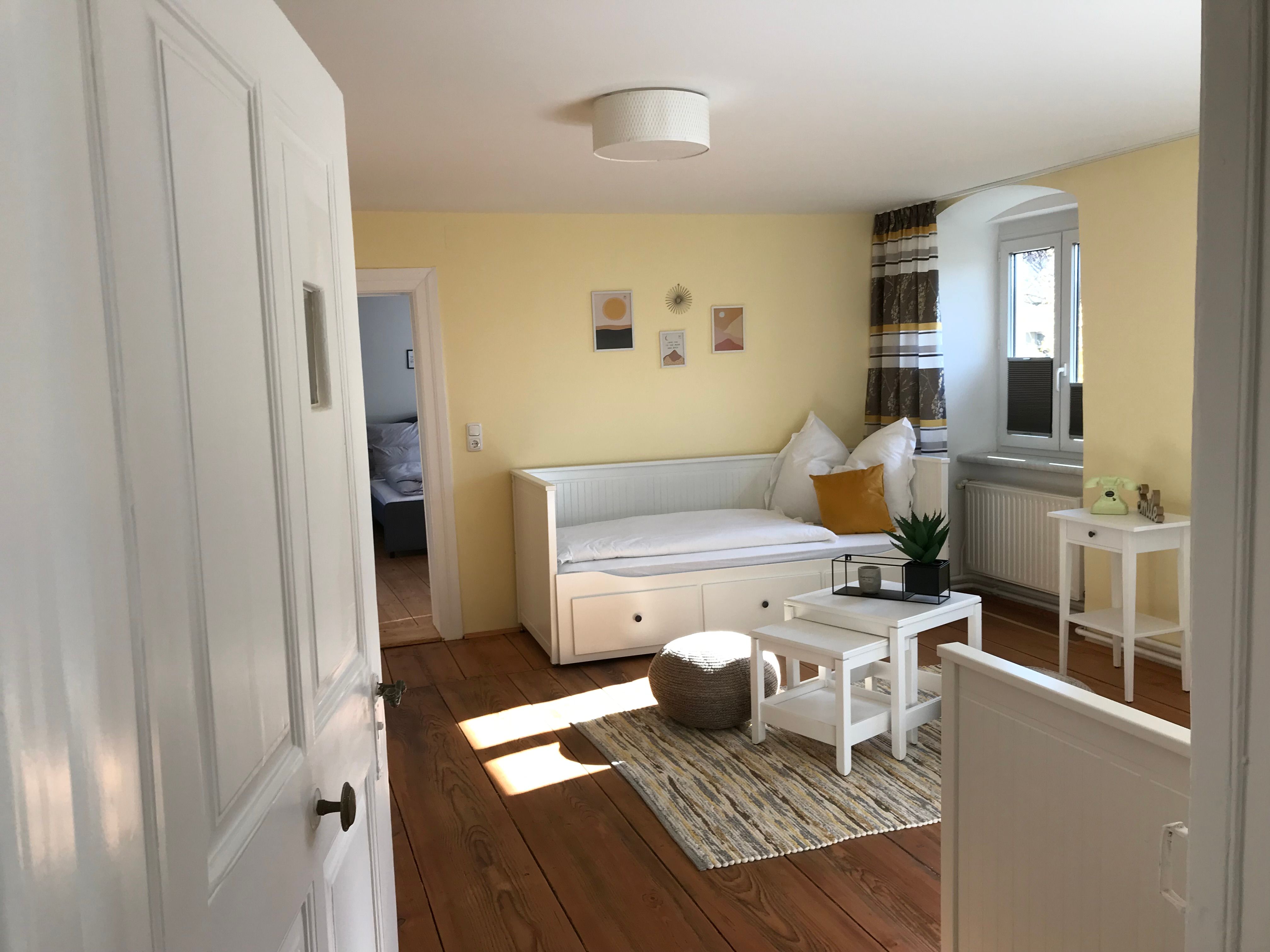 Világos szoba sárga falakkal, fehér nappali ággyal, kis asztallal és növényekkel.