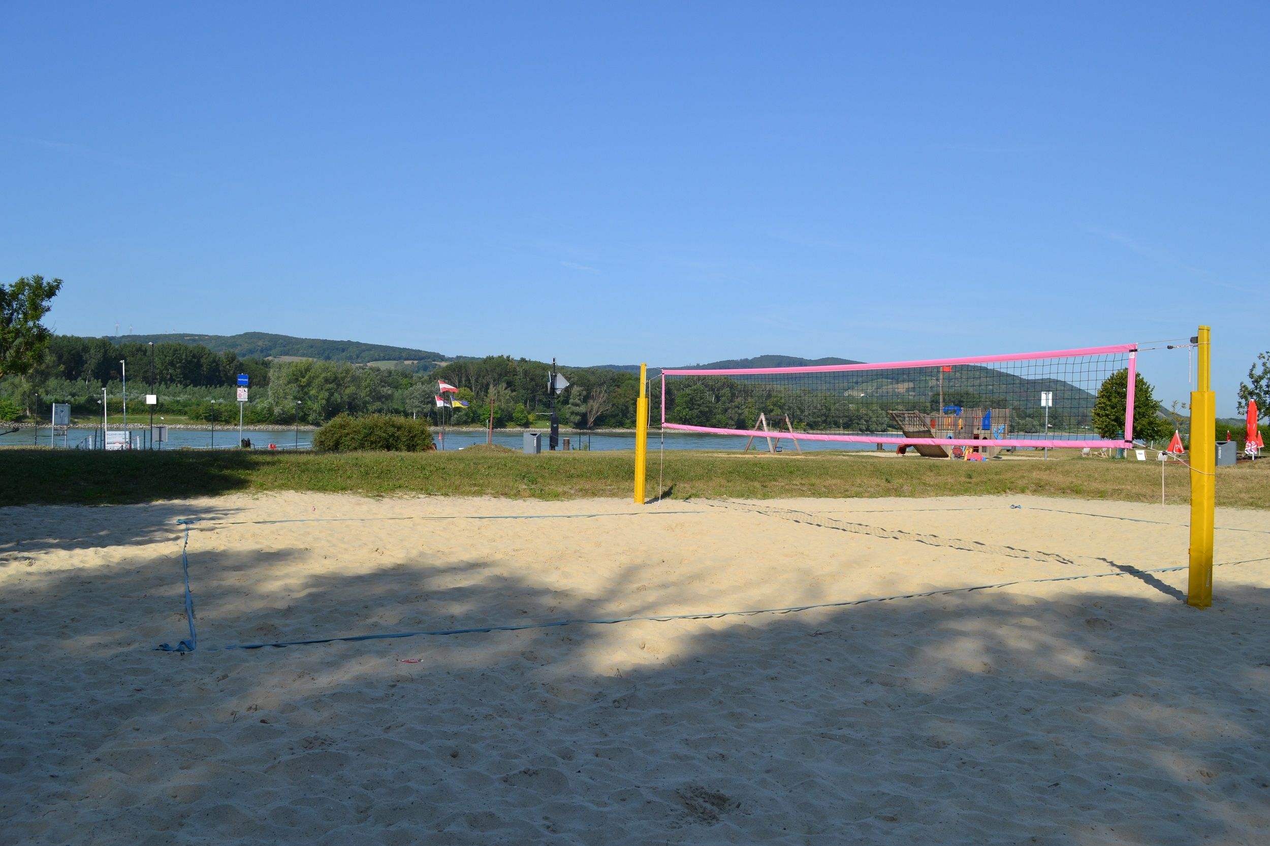 Egy üres strandröplabdapálya sárga oszlopokkal és rózsaszín hálóval, a háttérben egy folyó és zöld dombok előtt.