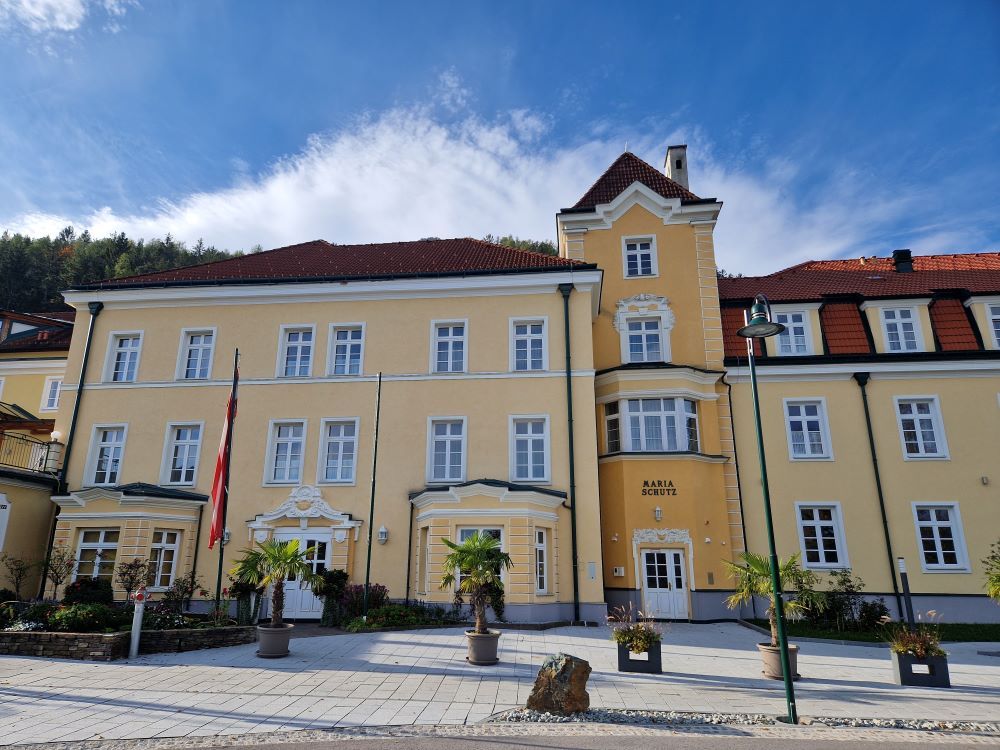 Sárga épület piros tetővel és több ablakkal, növényekkel és zászlókkal körülvéve.