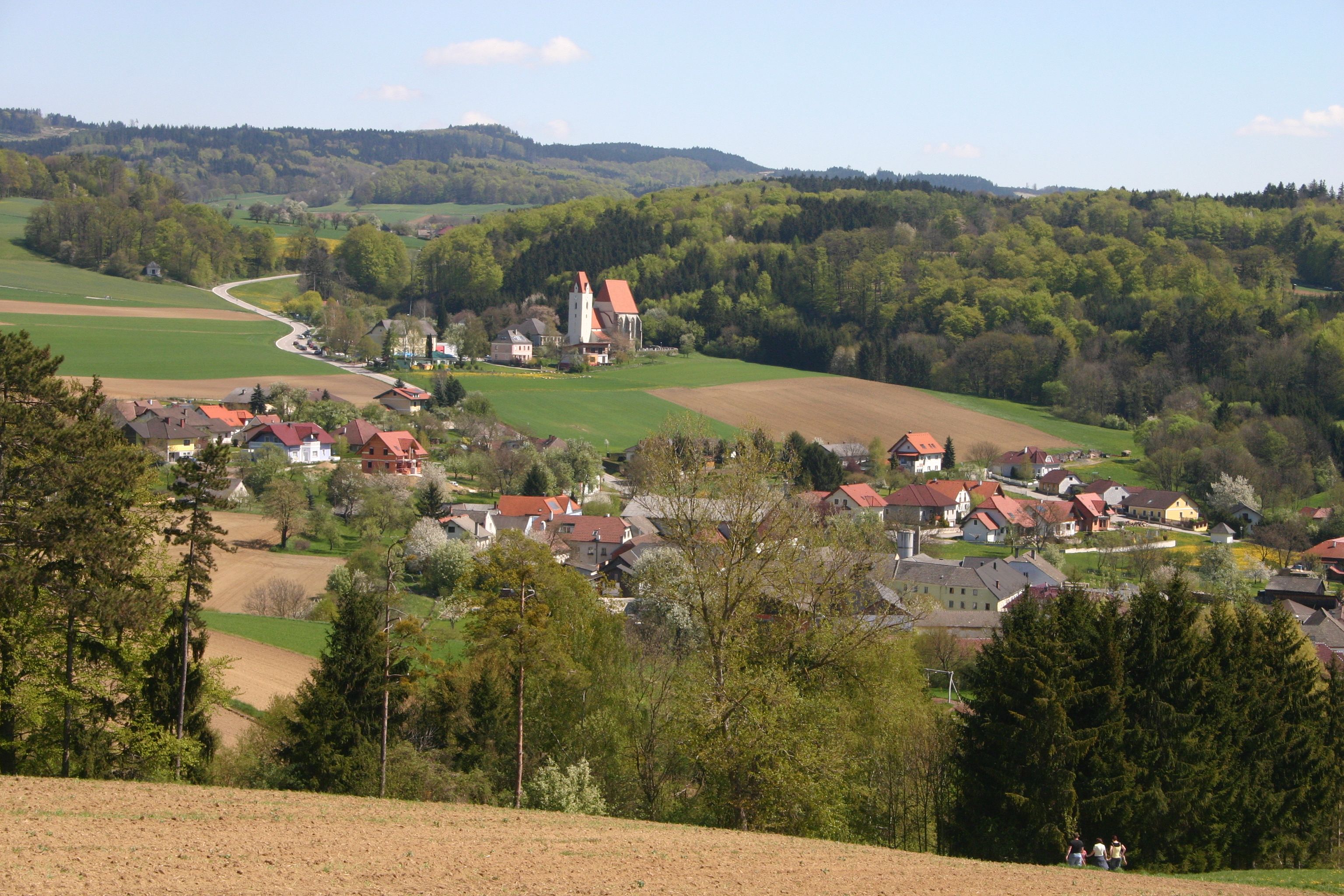 Tájkép faluval, templommal és dombokkal a háttérben.