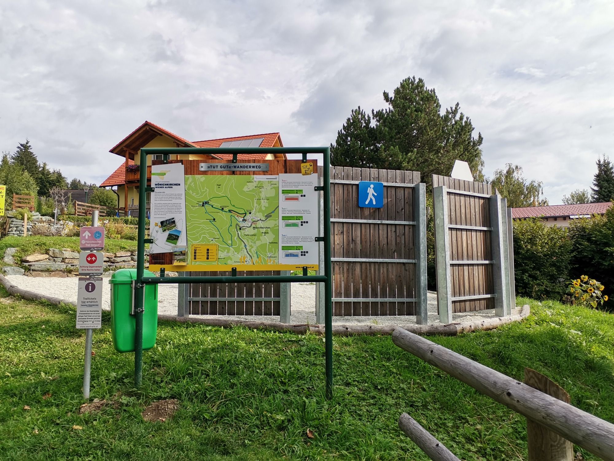 Túraútvonal információs tábla térképével és táblákkal egy réten, fákkal és egy épülettel a háttérben.
