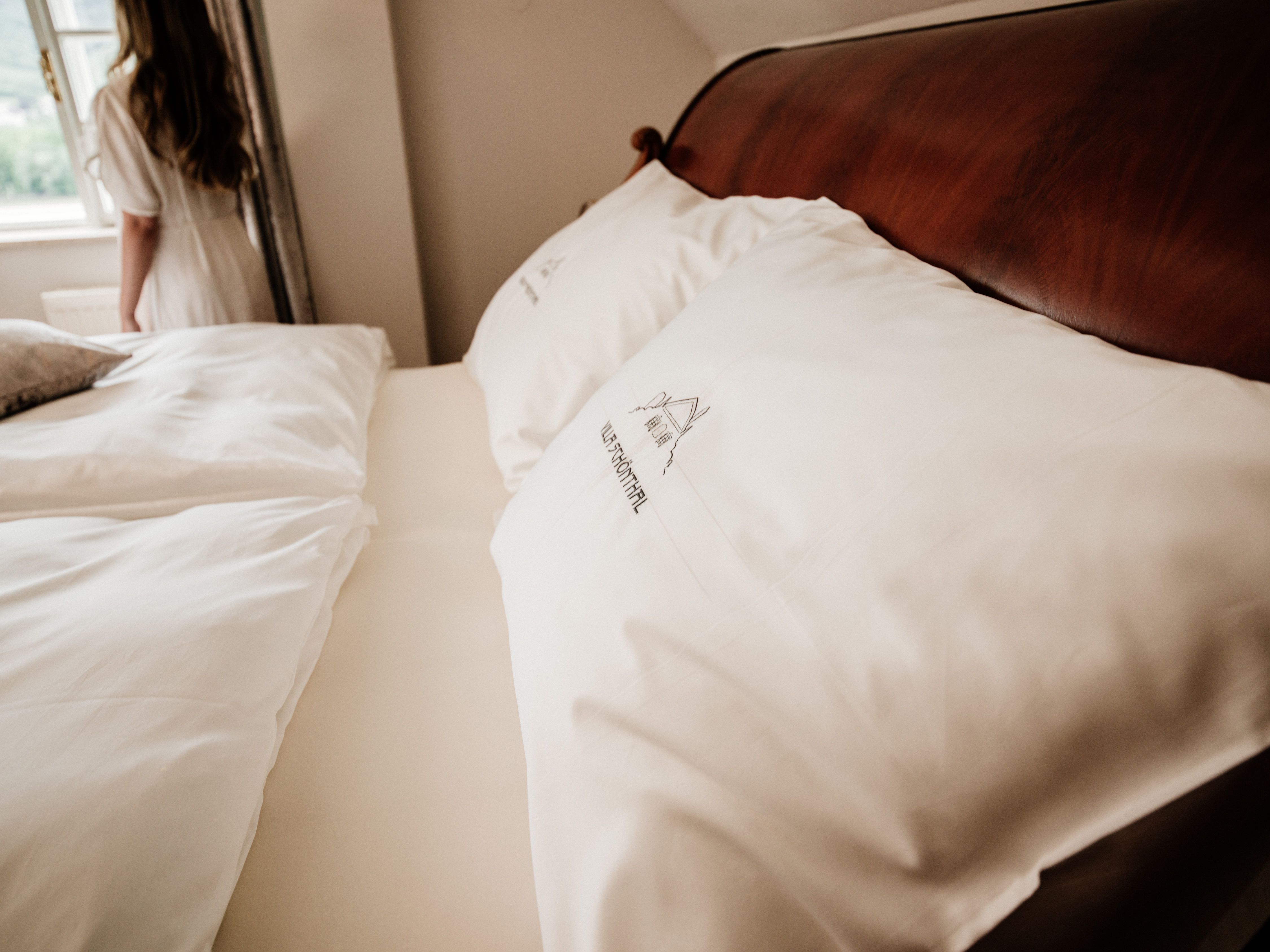 Közelkép egy ágyról, fehér párnákkal és ágyneművel, a háttérben egy nő az ablaknál.