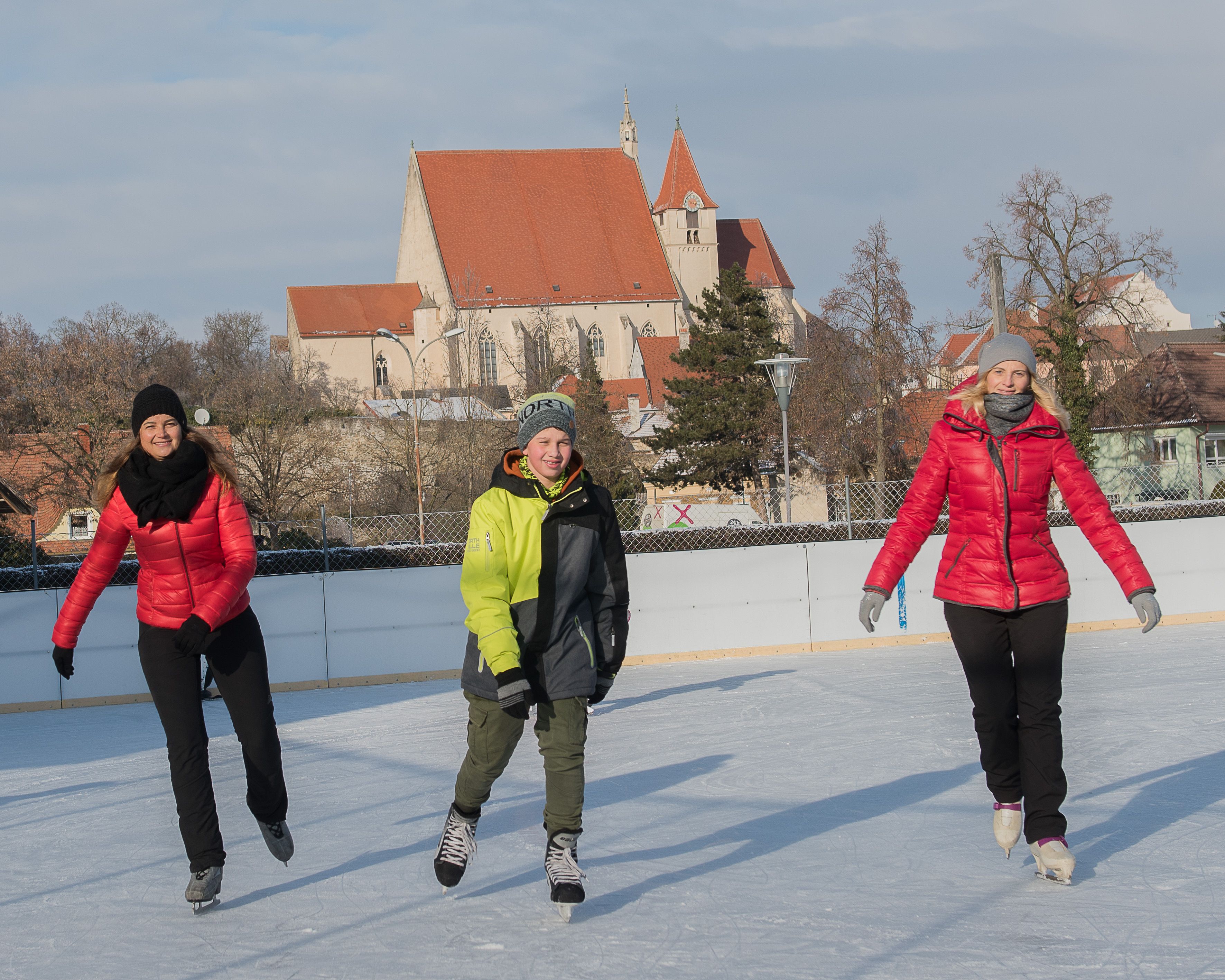 Három ember korcsolyázik egy jégpályán, a háttérben egy templom.
