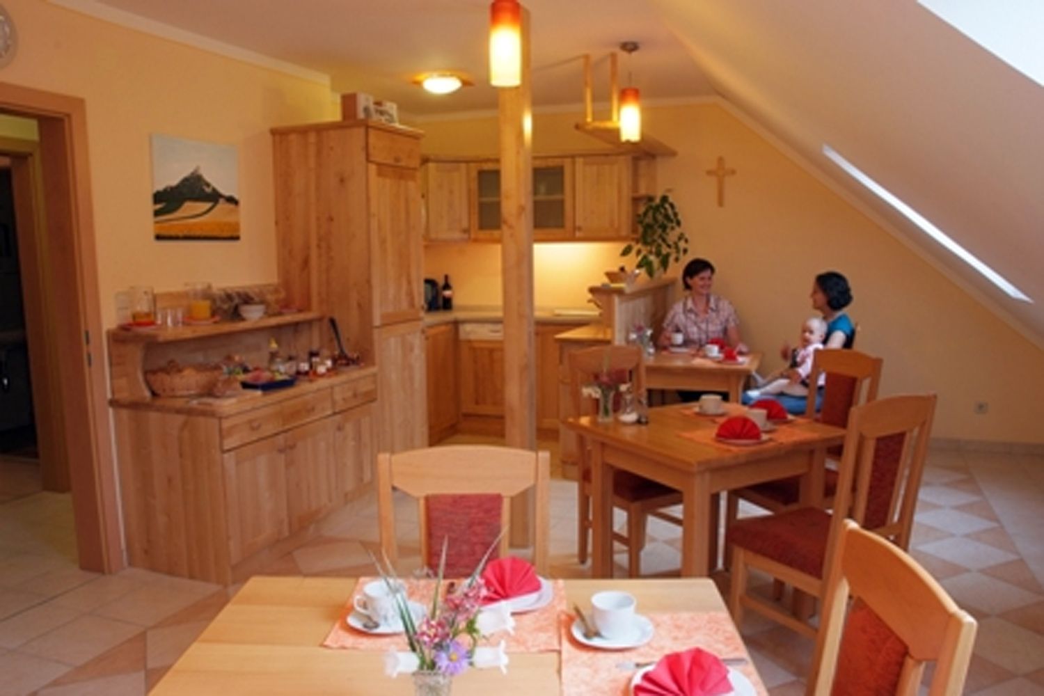 Hangulatos reggelizőszoba, fabútorokkal, terített asztalokkal, két beszélgető emberrel. Az egyikük ölében egy gyermek ül.