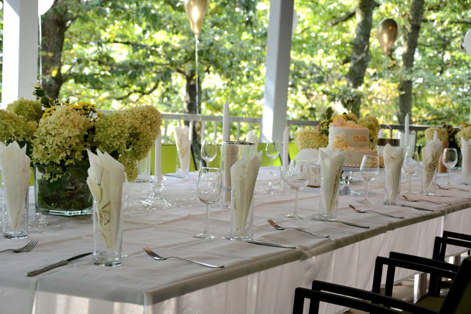 Ünnepélyesen megterített fehér asztal a teraszon, virágokkal és gyertyákkal.