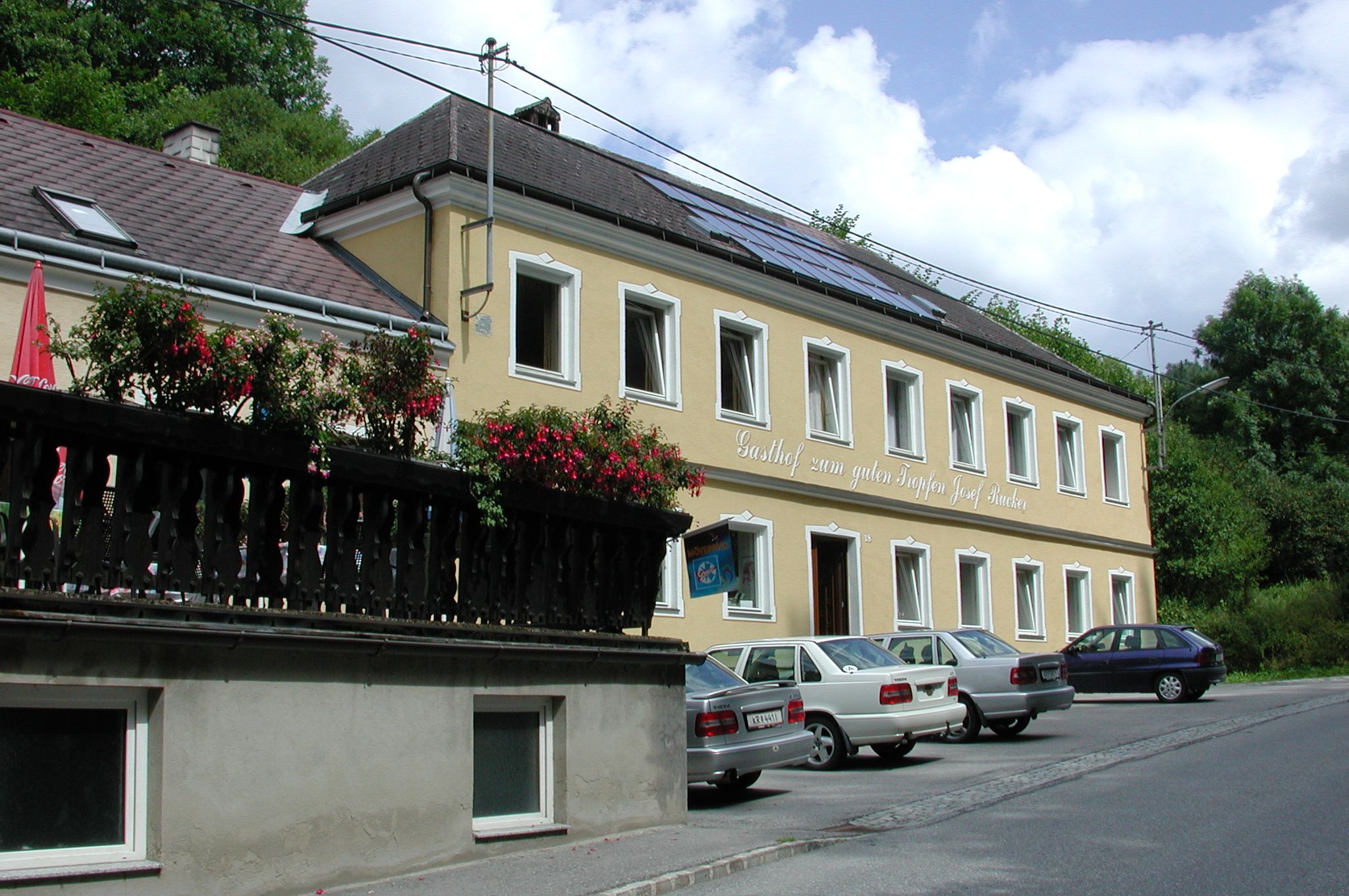 "Gasthof zum guten Tropfen Josef Rucker" feliratú sárga épület, előtte parkoló autók, háttérben kék égbolt.
