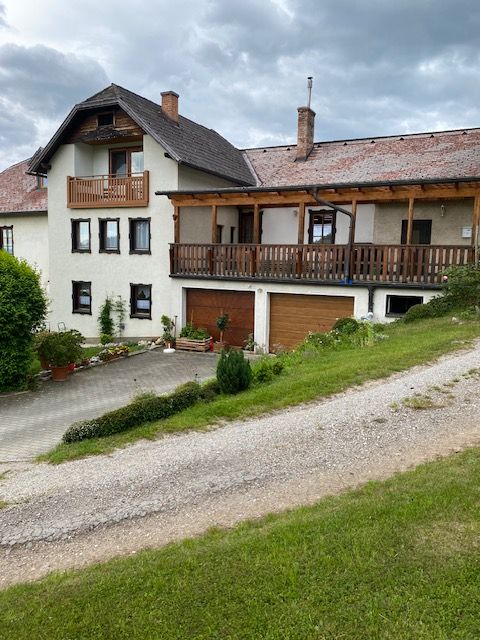 Egy kétszintes ház erkéllyel és garázzsal, zöld vidékkel és felhős égboltal körülvéve.
