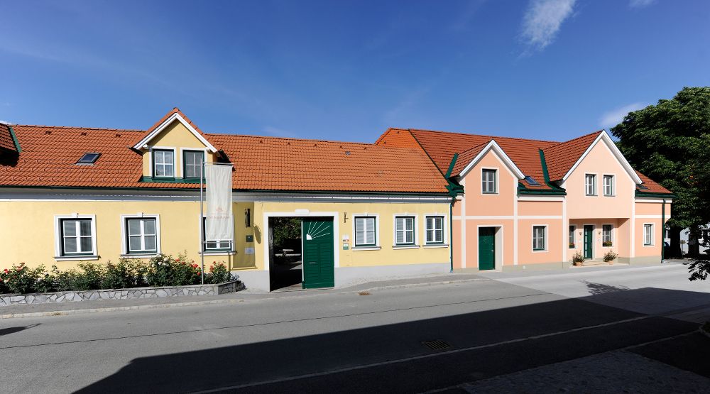 Egy hosszú, egyemeletes ház, sárga és rózsaszín homlokzattal, zöld redőnyökkel és piros cseréptetővel, kék ég alatt.