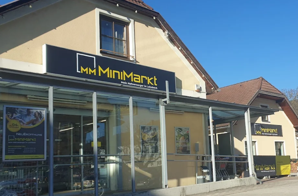 Egy MiniMarkt üzlet külső nézete üveghomlokzattal és reklámplakátokkal.