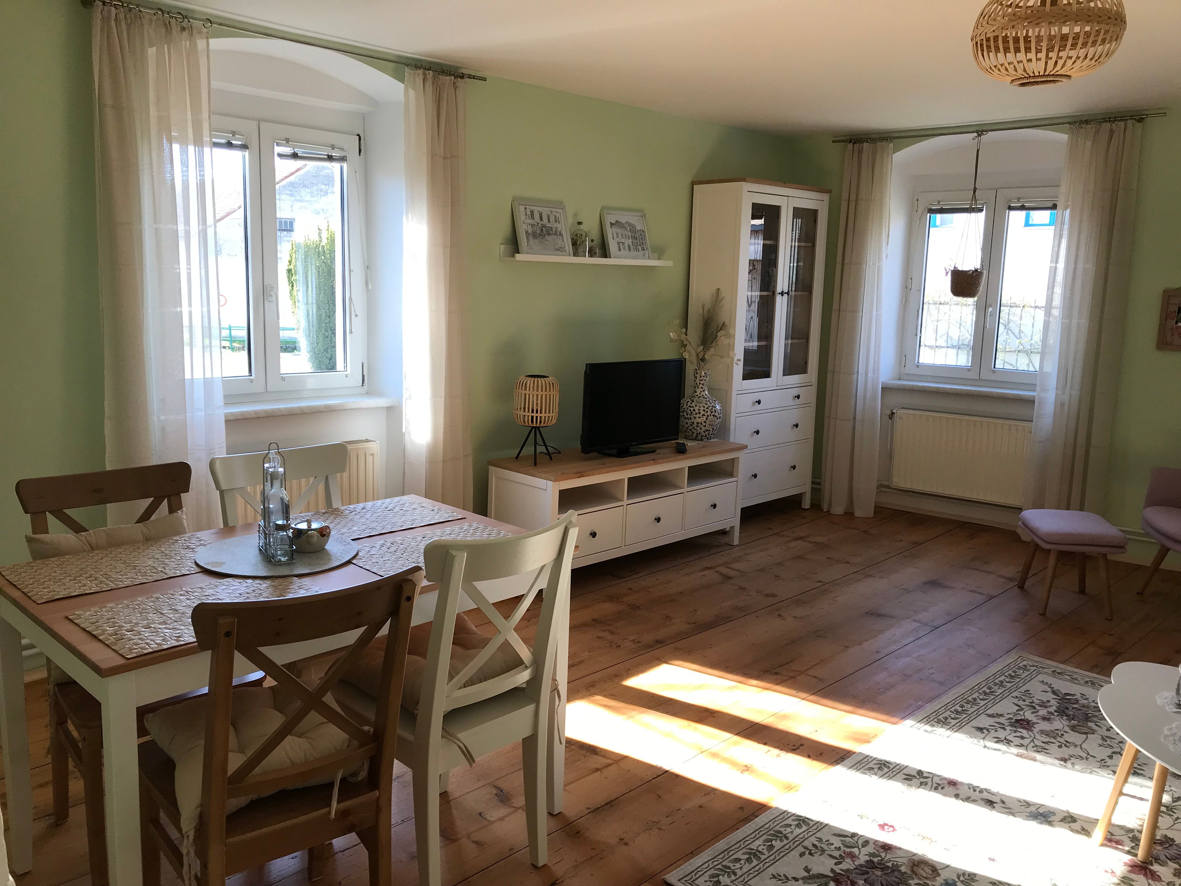 Világos szoba étkezőasztallal, székekkel, TV-szekrénnyel és nagy ablakokkal.