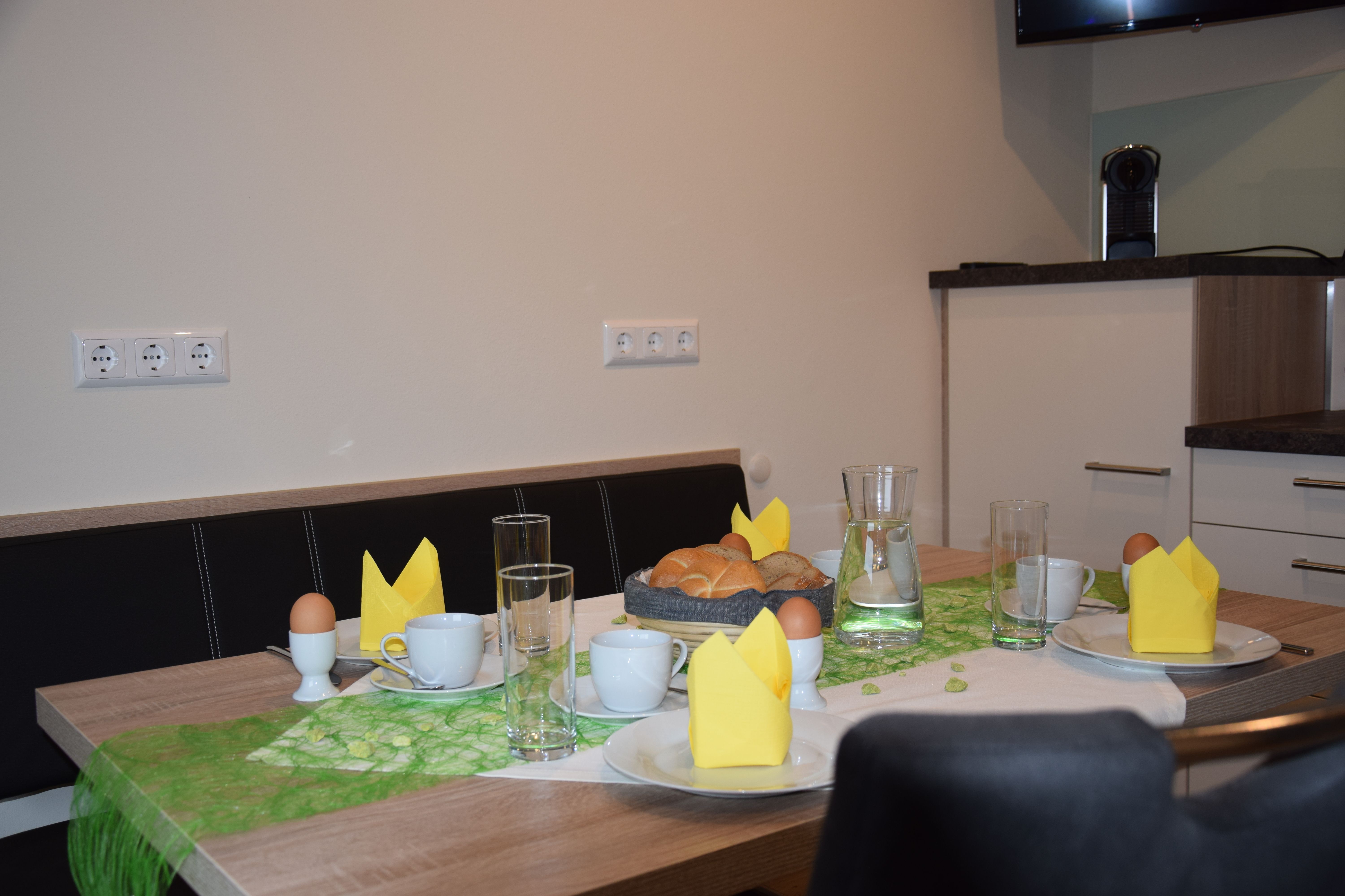 Reggelizőasztal zsemlével, tojással, csészékkel és sárga szalvétával.