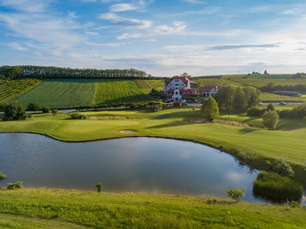 Tájképi felvétel egy golfpályáról, a háttérben tóval és klubházzal.