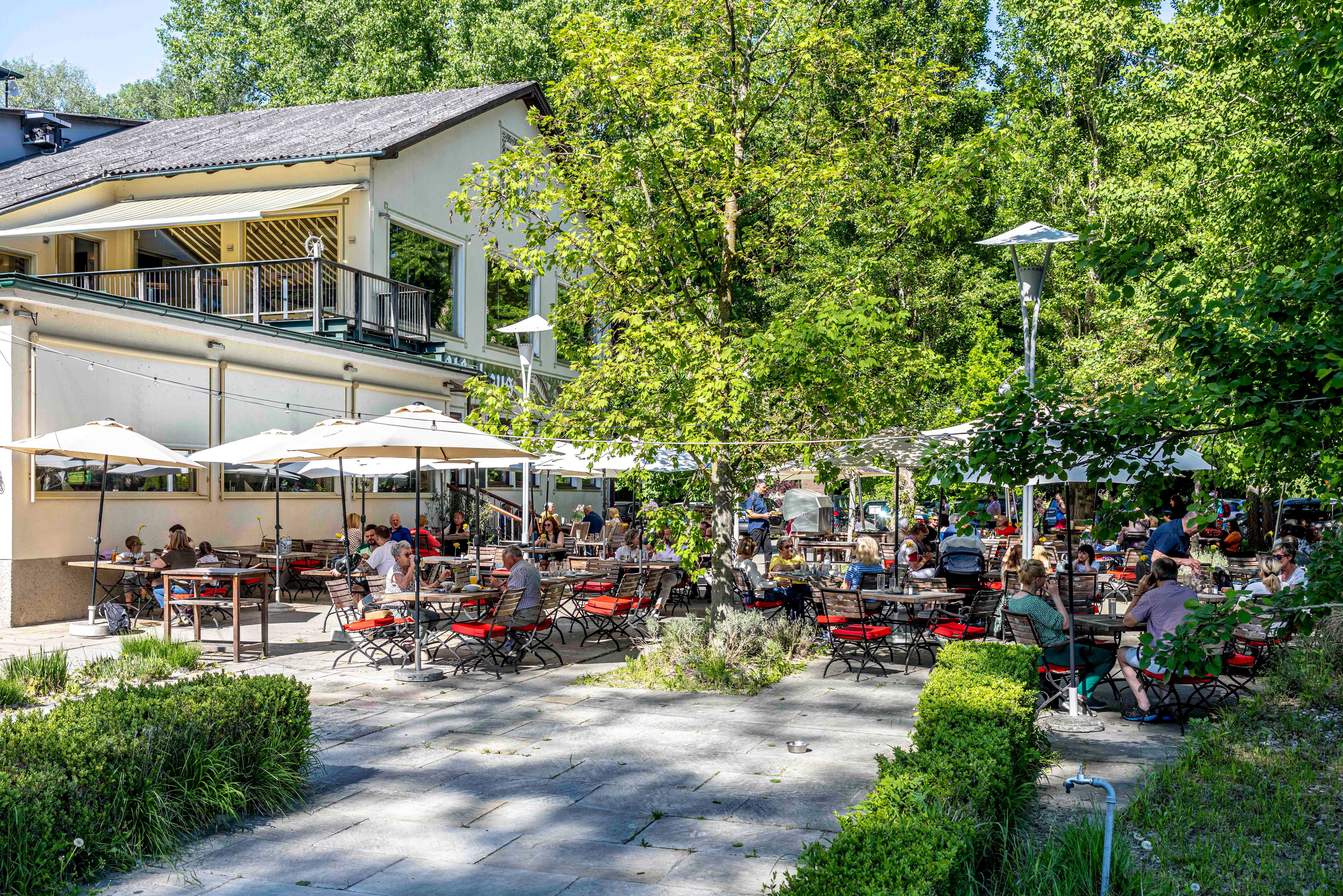 Egy vendégkert, ahol sok ember ül az asztaloknál, napernyők alatt, körülötte fák és egy épület a háttérben.
