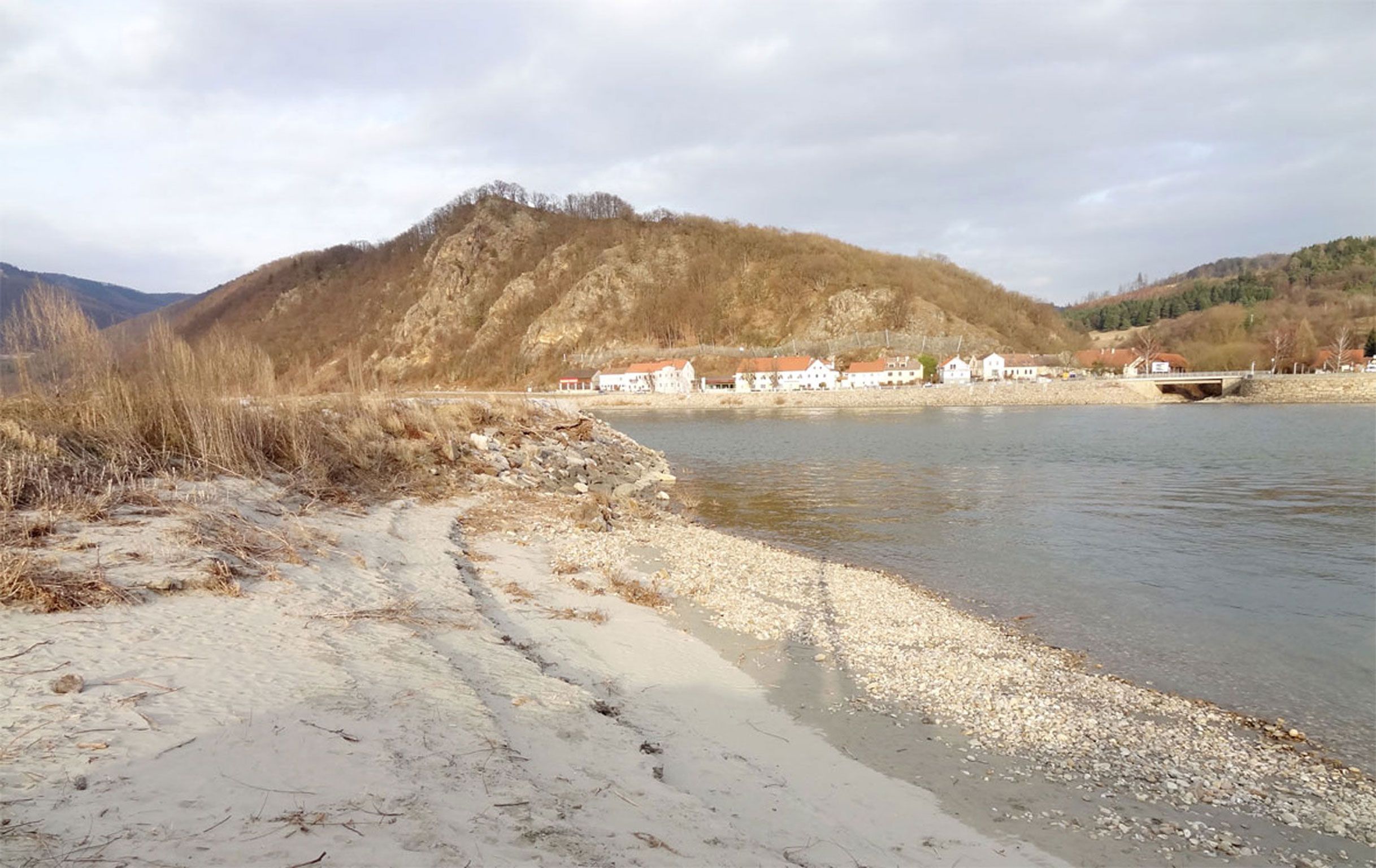 Csendes, kavicsos strand, a háttérben egy falu és erdős dombok.